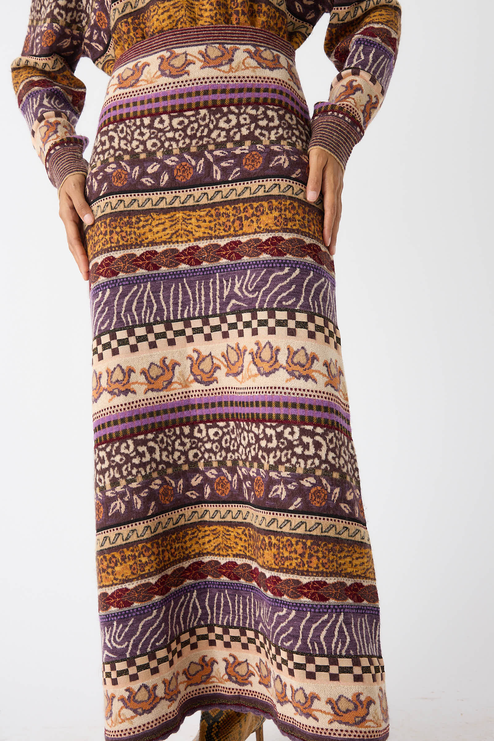 Close-up of a person wearing the Ulla Johnson Helene Skirt in Woodland, along with a matching sweater featuring Fair Isle motifs in shades of purple, orange, red, and white. Hands rest gently on either side of the long patterned knit skirt.