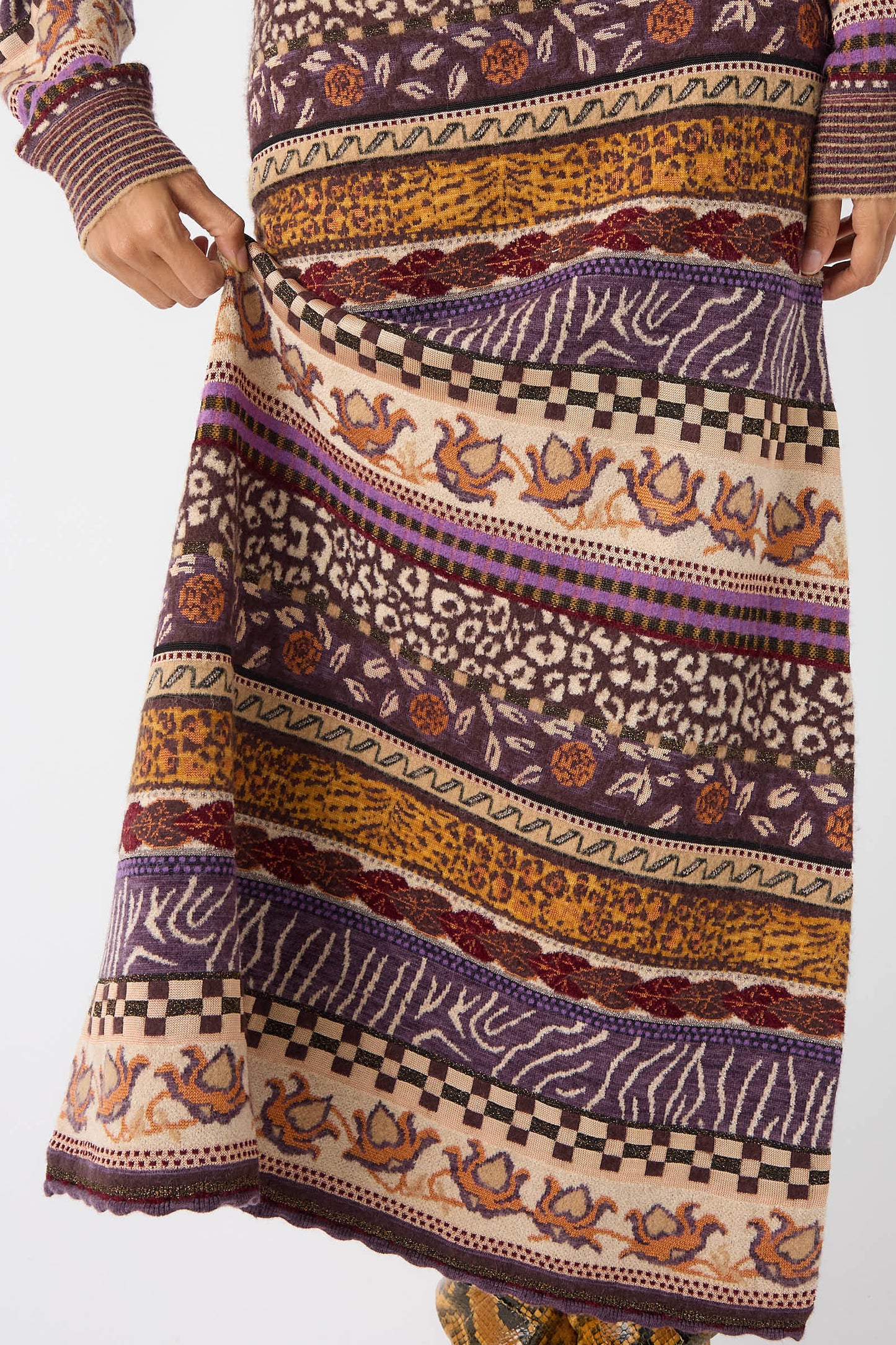 A close-up of a person holding the hem of the Helene Skirt in Woodland by Ulla Johnson, showcasing its multicolored, patterned knit design featuring stripes, florals, and geometric shapes inspired by woodland colors.