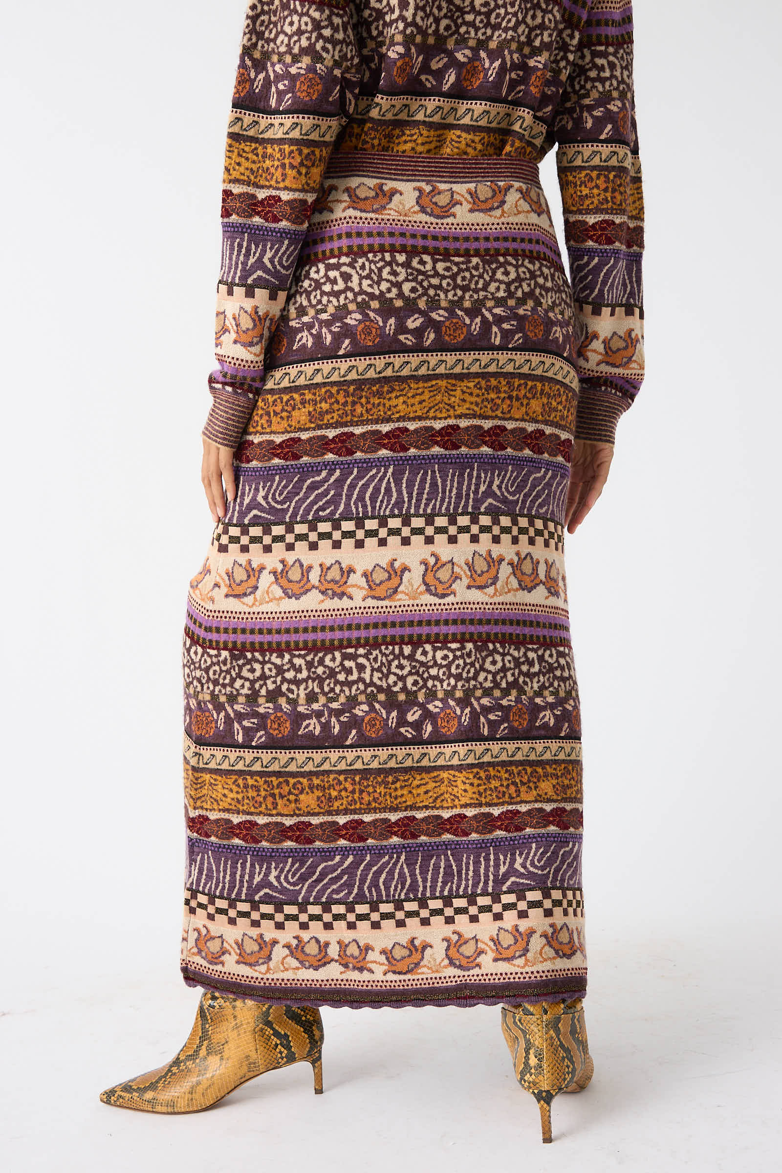 A person wearing the Helene Skirt in Woodland by Ulla Johnson, along with a matching top, stands facing away. The long knit skirt showcases horizontal stripes with Fair Isle motifs combined with animal and abstract designs, all in a harmonious mix of woodland colors.