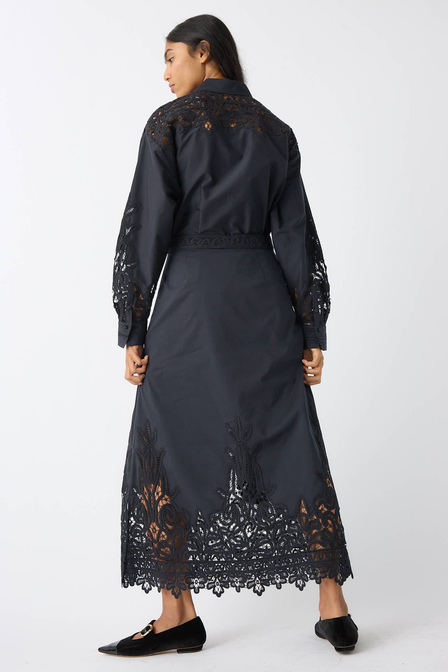 A person stands facing away, wearing the Ulla Johnson Karlyn Skirt in Noir with Battenberg lace detailing, paired with black flats. They have long dark hair.