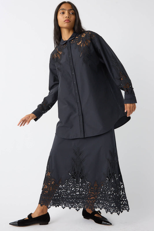 A person wears a black long-sleeved shirt and the Ulla Johnson Karlyn Skirt in Noir, which features Victorian-inspired lace detailing. They are also wearing black flats and standing against a plain background.