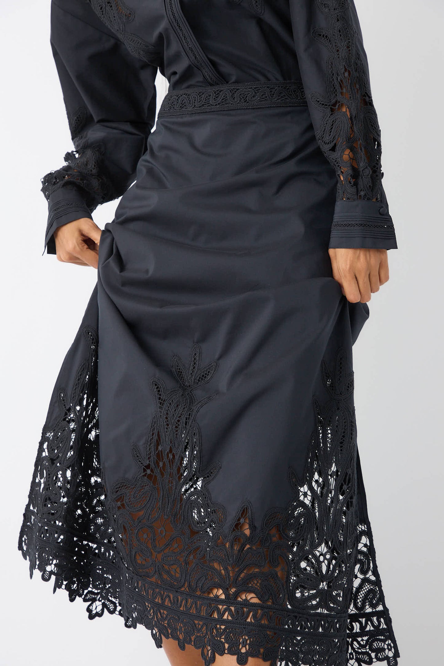 A person wearing the Ulla Johnson Karlyn Skirt in Noir holds the skirt slightly lifted, revealing detailed Battenberg lace along the hem and sleeves, adding an elegant touch reminiscent of a Victorian-inspired design.