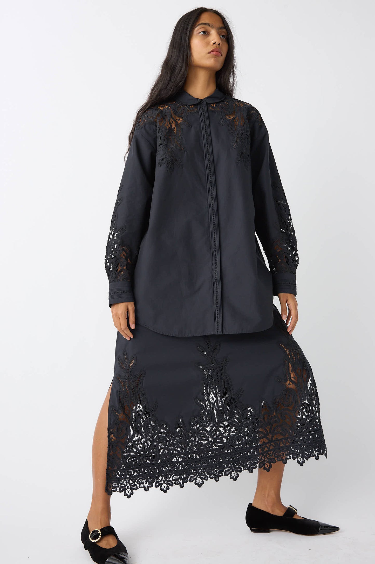 A woman stands facing forward, wearing a black blouse and the Ulla Johnson Karlyn Skirt in Noir, which features intricate Battenberg lace detailing on the sleeves, chest, and hem. She completes the outfit with matching black shoes.