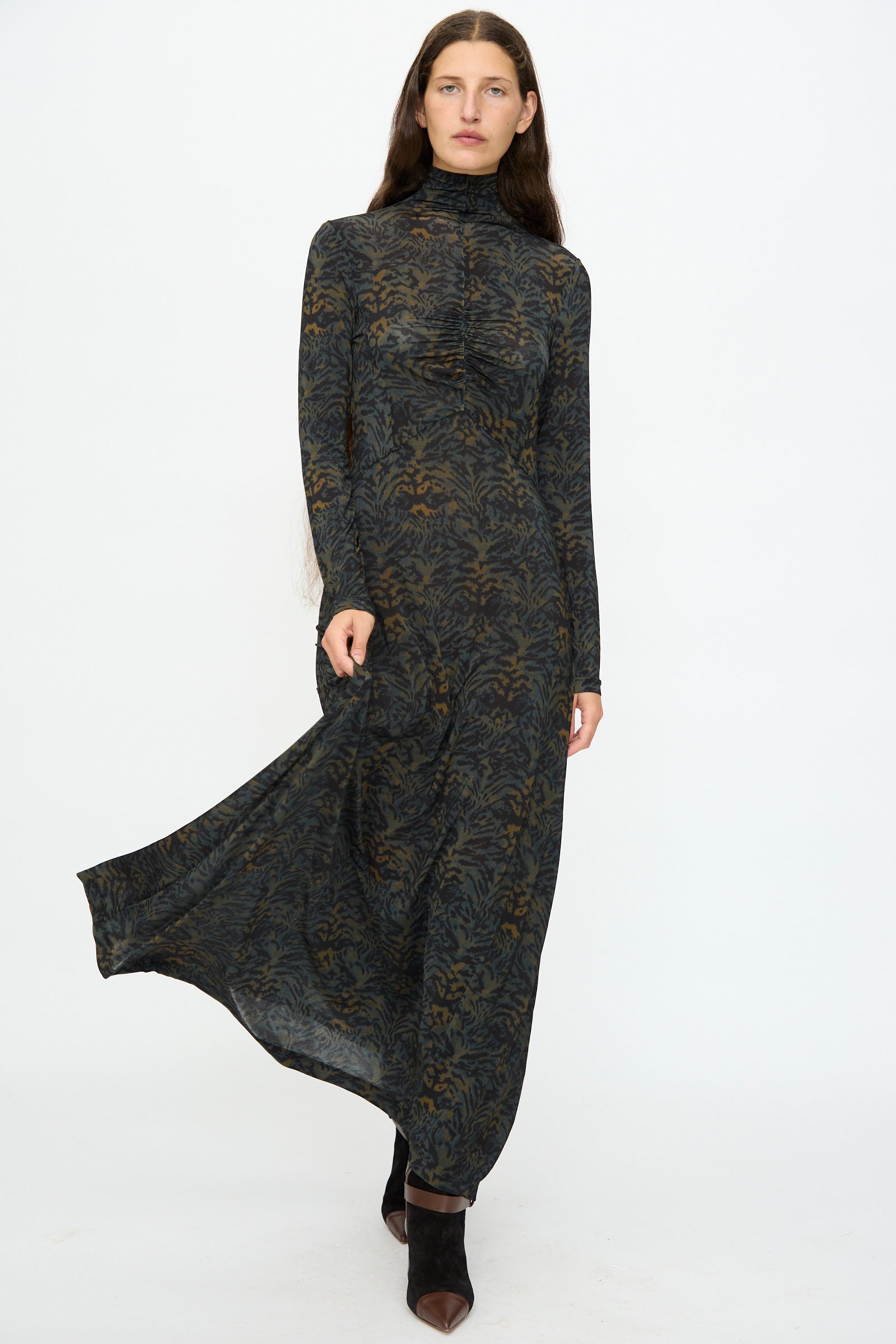 A woman stands against a plain background wearing the Ulla Johnson Koa Dress in Desert Jasper, a slim fit maxi with long sleeves and high neck, featuring a dark ikat animal print. She pairs it with black pointed-toe shoes and lifts the hem slightly.