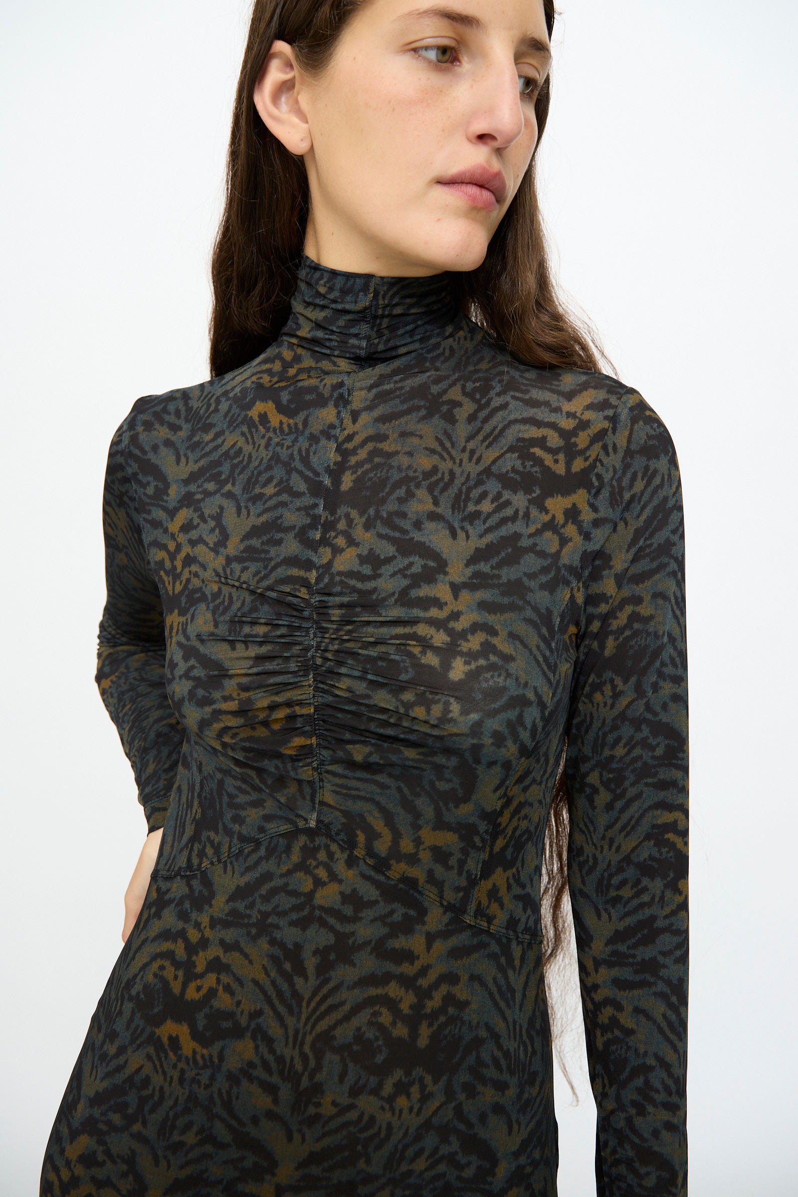 A person wears the Ulla Johnson Koa Dress in Desert Jasper—a slim fit, long sleeve maxi with a high neck and ruched chest detail in a dark ikat animal print—standing against a plain white background.
