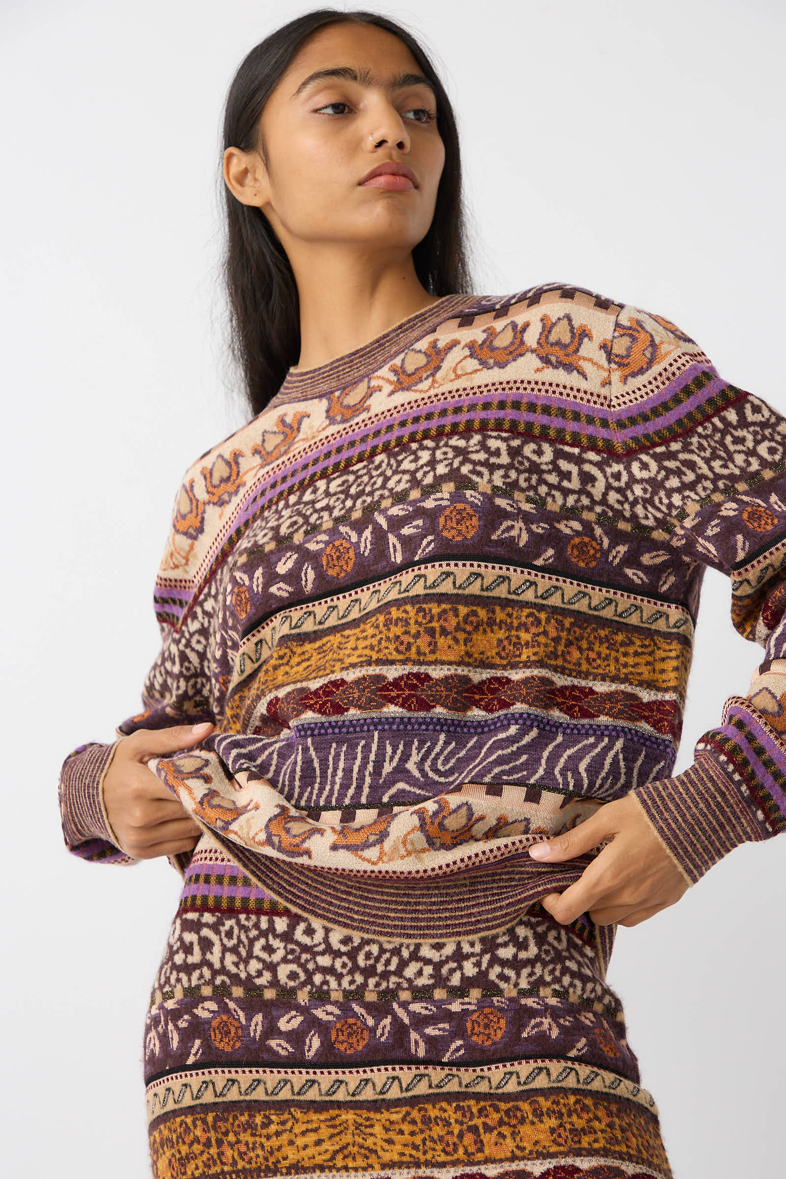 A person wearing the Ulla Johnson Lara Pullover in Woodland, a colorful sweater with a mix of geometric and animal print designs, featuring Fair Isle motifs in mid-weight plush yarn, poses against a plain background.