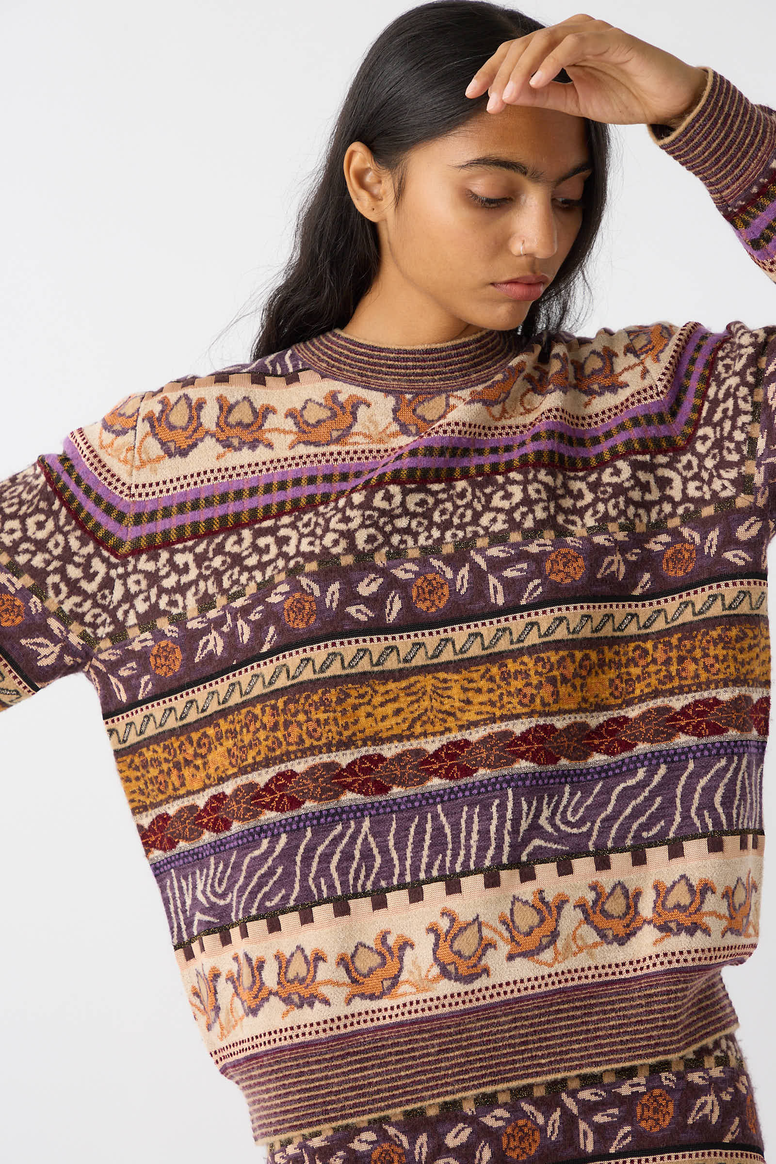 A person with long hair dons the Ulla Johnson Lara Pullover in Woodland, featuring various geometric and floral designs in earth tones, purple, and orange, while looking downward and raising one arm.