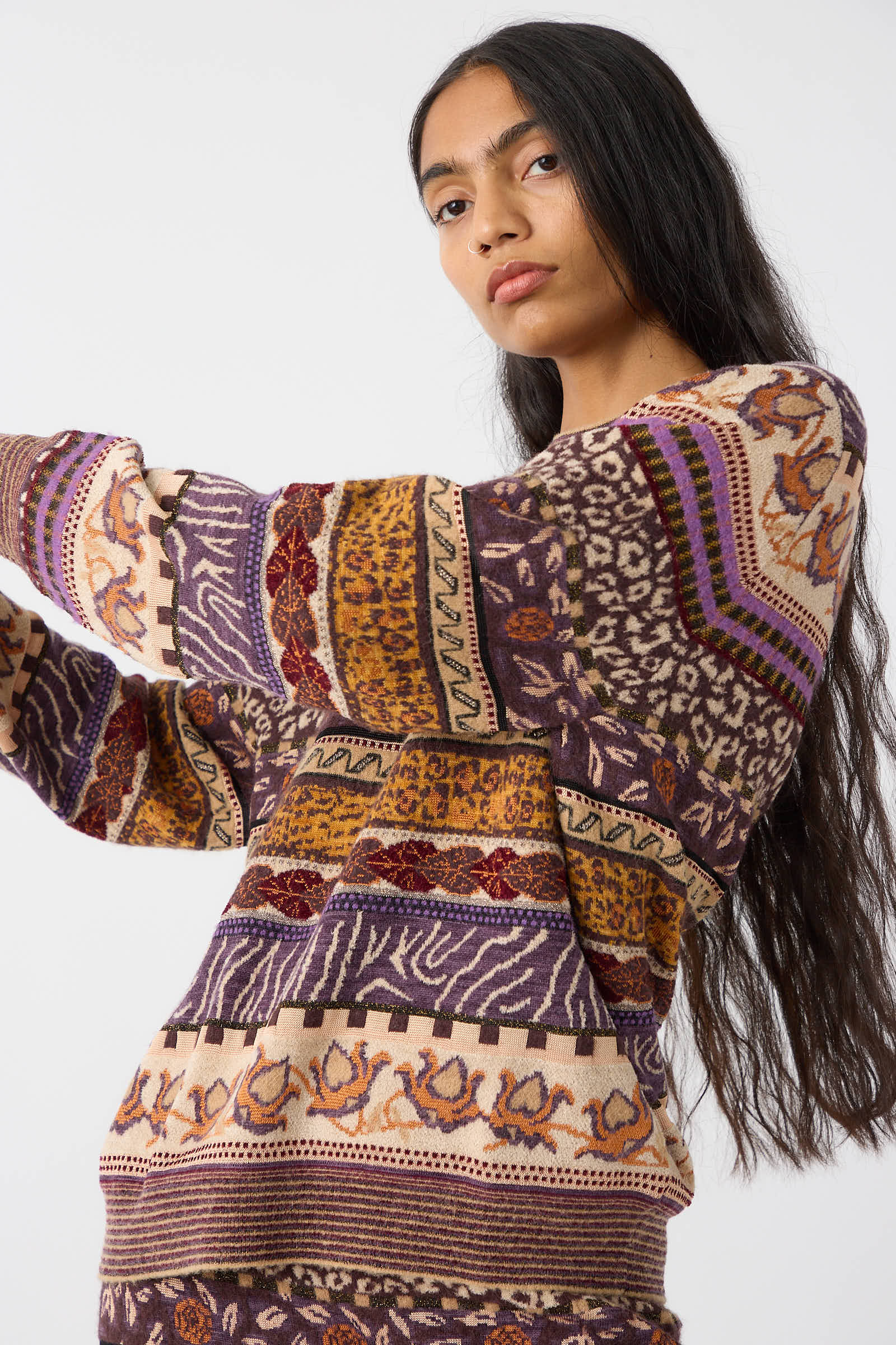 A person with long dark hair wears the Ulla Johnson Lara Pullover in Woodland, showcasing colorful, patterned designs in hues of purple, orange, and beige while looking at the camera. The mid-weight plush yarn adds a cozy feel to the Fair Isle motifs adorning the sweater.