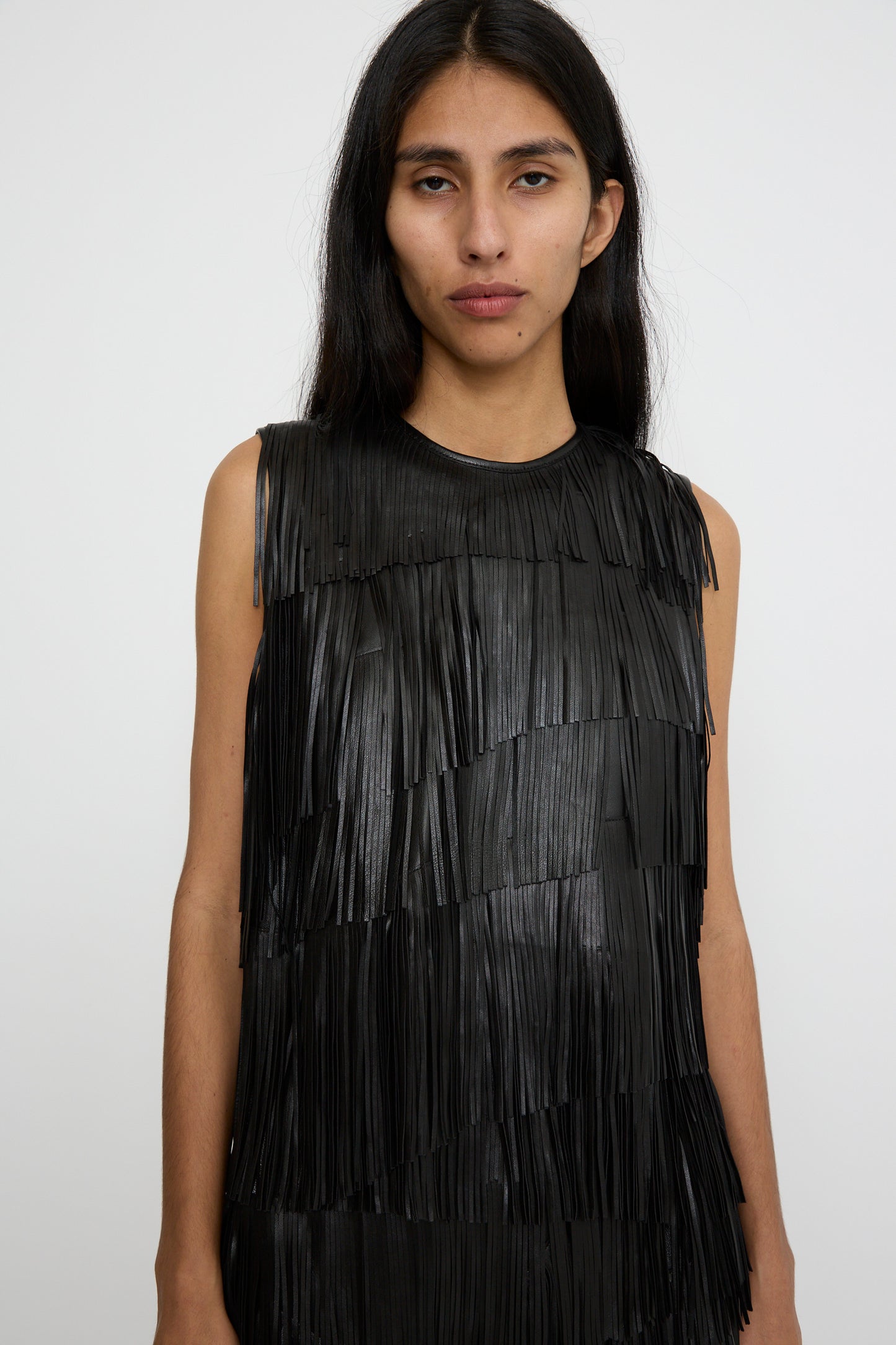 A person stands forward against a plain white background, wearing the Leonie Top in Noir by Ulla Johnson, featuring a sleeveless black design with raw-edge fringe.