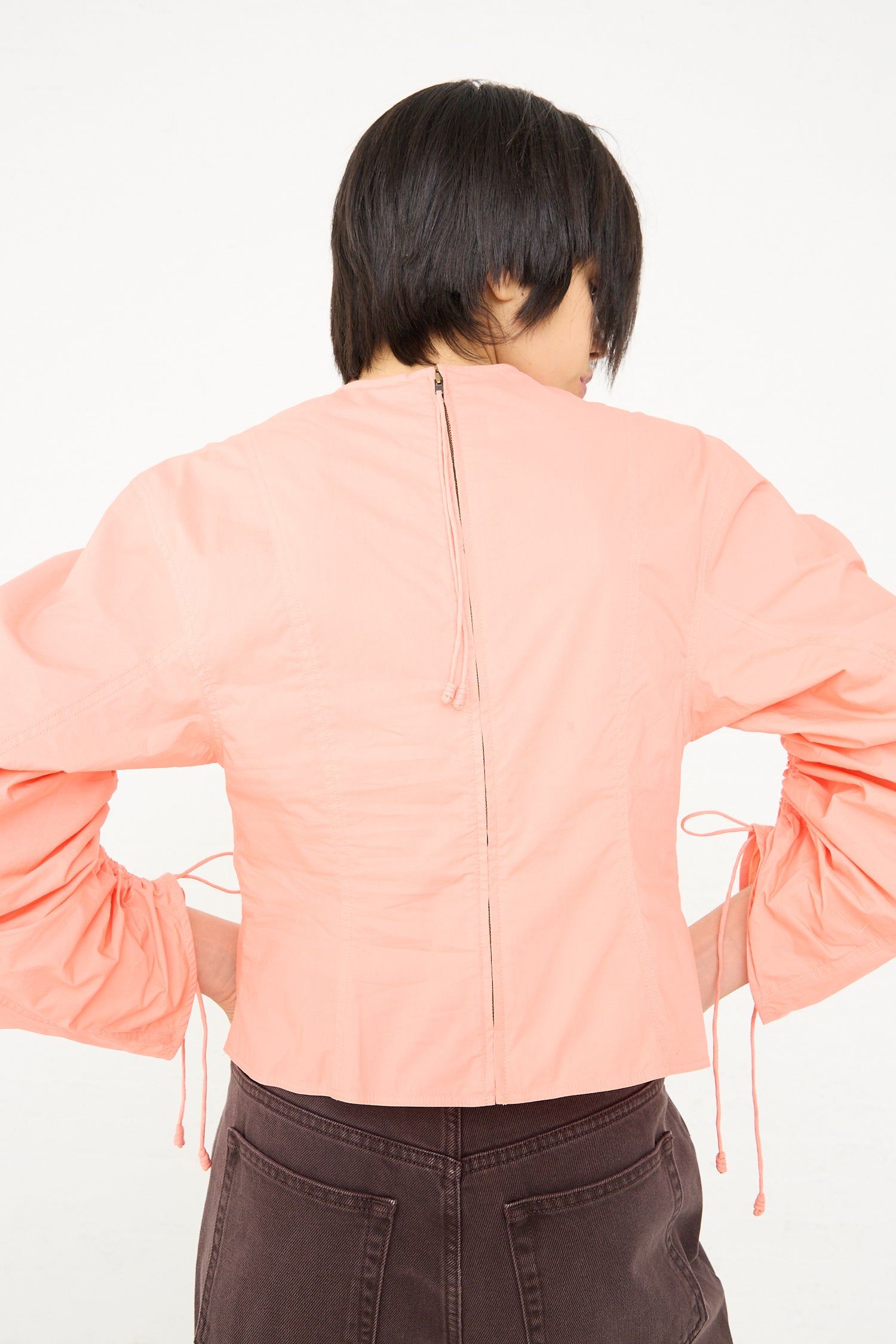 Woman stands with her back to the camera, wearing a Ulla Johnson Lorna Blouse in Peony with ruched sleeves and brown pants.