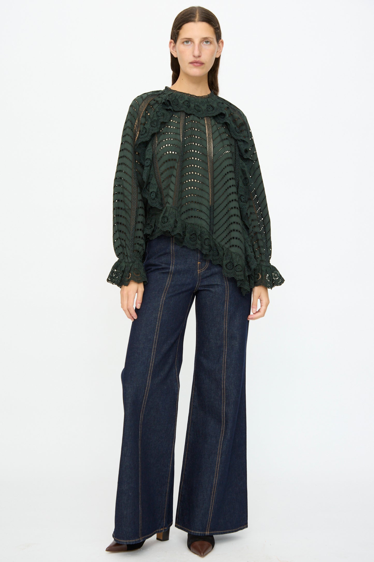 A person stands facing forward, wearing the Ulla Johnson Marcella Blouse in Cypress with scalloped ruffles and wide-leg dark denim jeans, paired with brown pointed-toe shoes against a plain background.