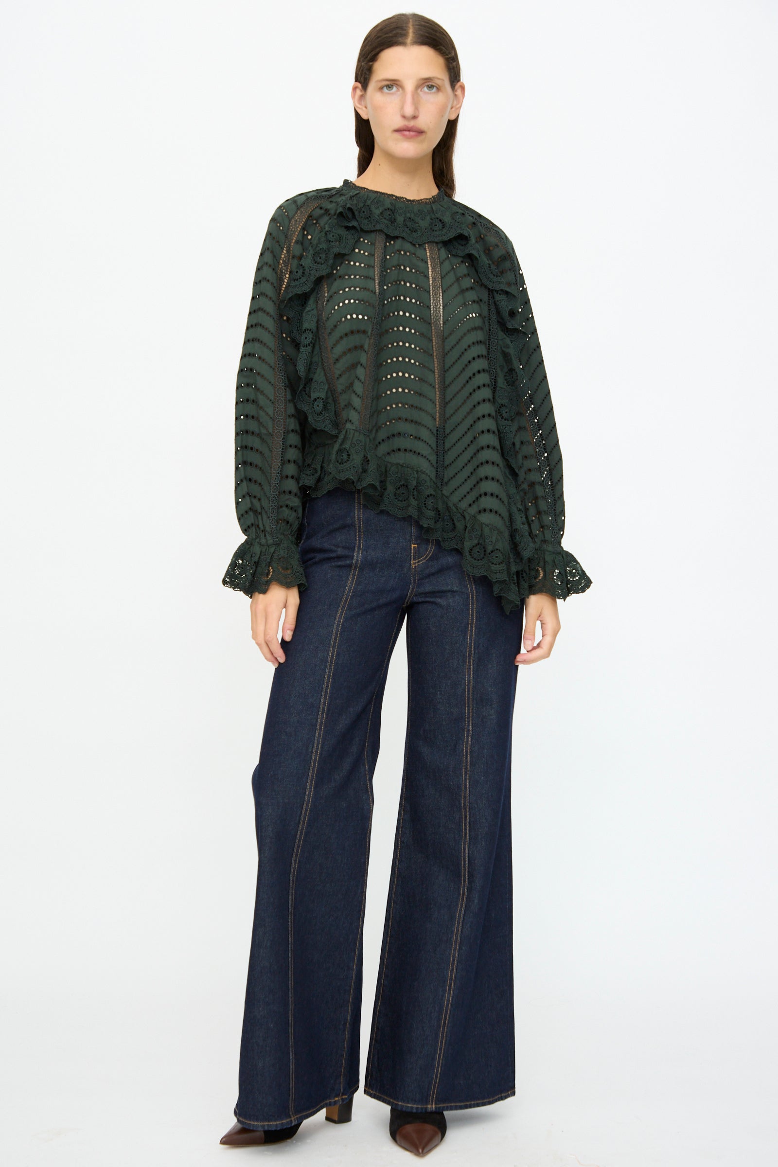 A person stands facing forward, wearing the Ulla Johnson Marcella Blouse in Cypress with scalloped ruffles and wide-leg dark denim jeans, paired with brown pointed-toe shoes against a plain background.