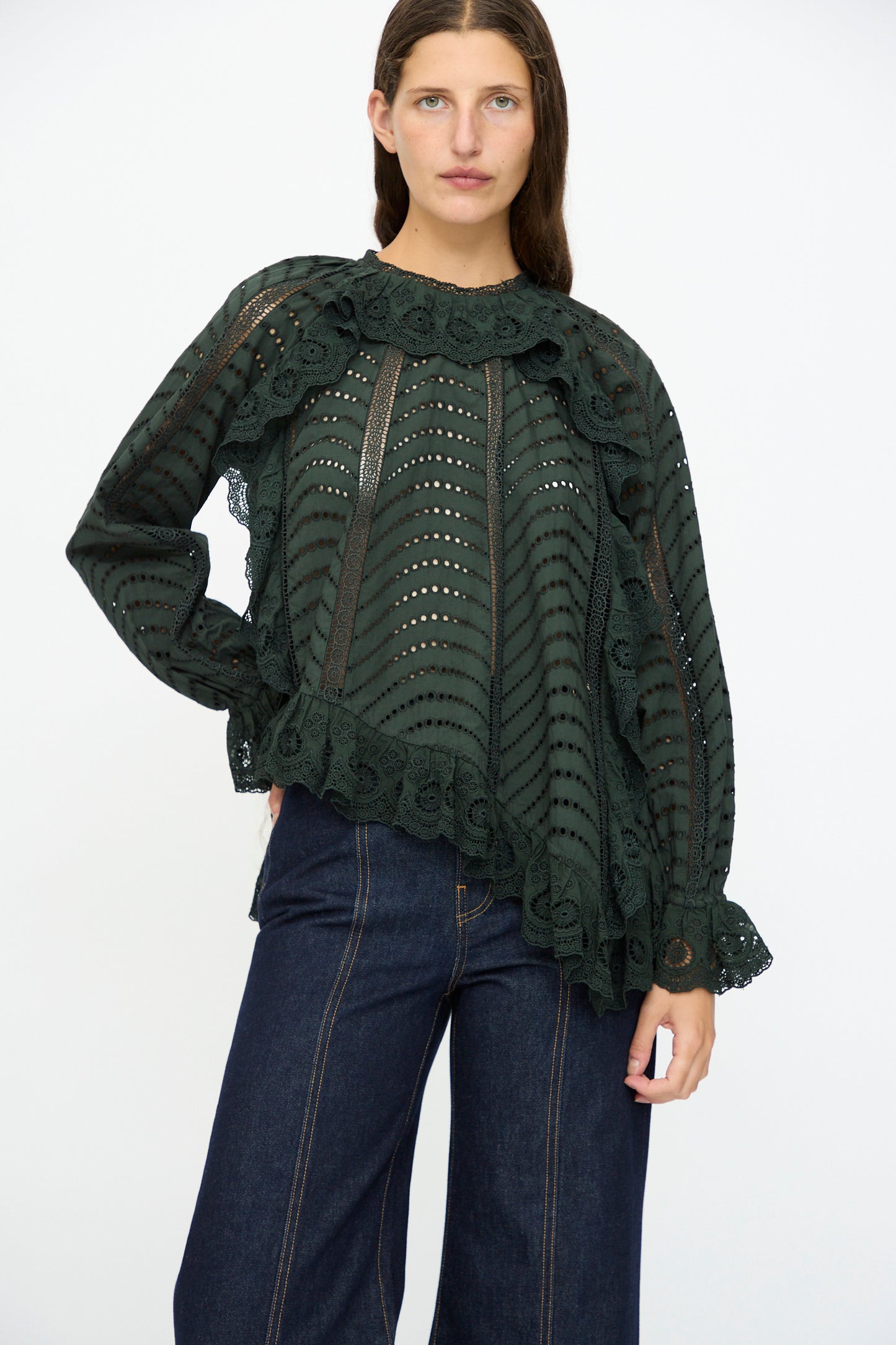A woman wears the Ulla Johnson Marcella Blouse in Cypress—a dark green, long sleeve top with scalloped ruffles—and wide-leg dark blue jeans, standing against a plain white background.