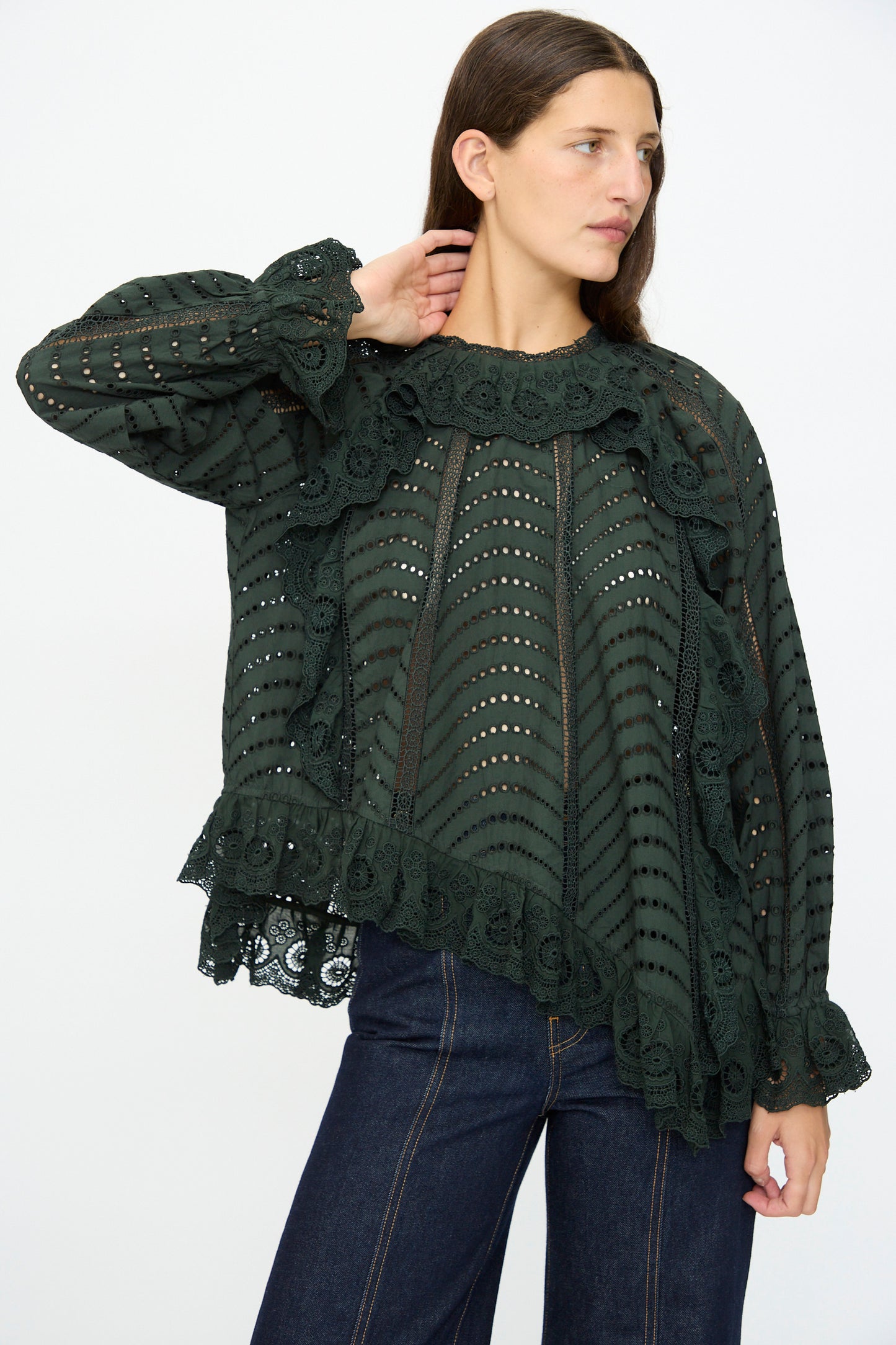 A woman wearing the Ulla Johnson Marcella Blouse in Cypress, a dark green cotton top with lace-trimmed eyelet details and scalloped ruffles, pairs it with dark denim jeans and stands with one hand touching her neck.