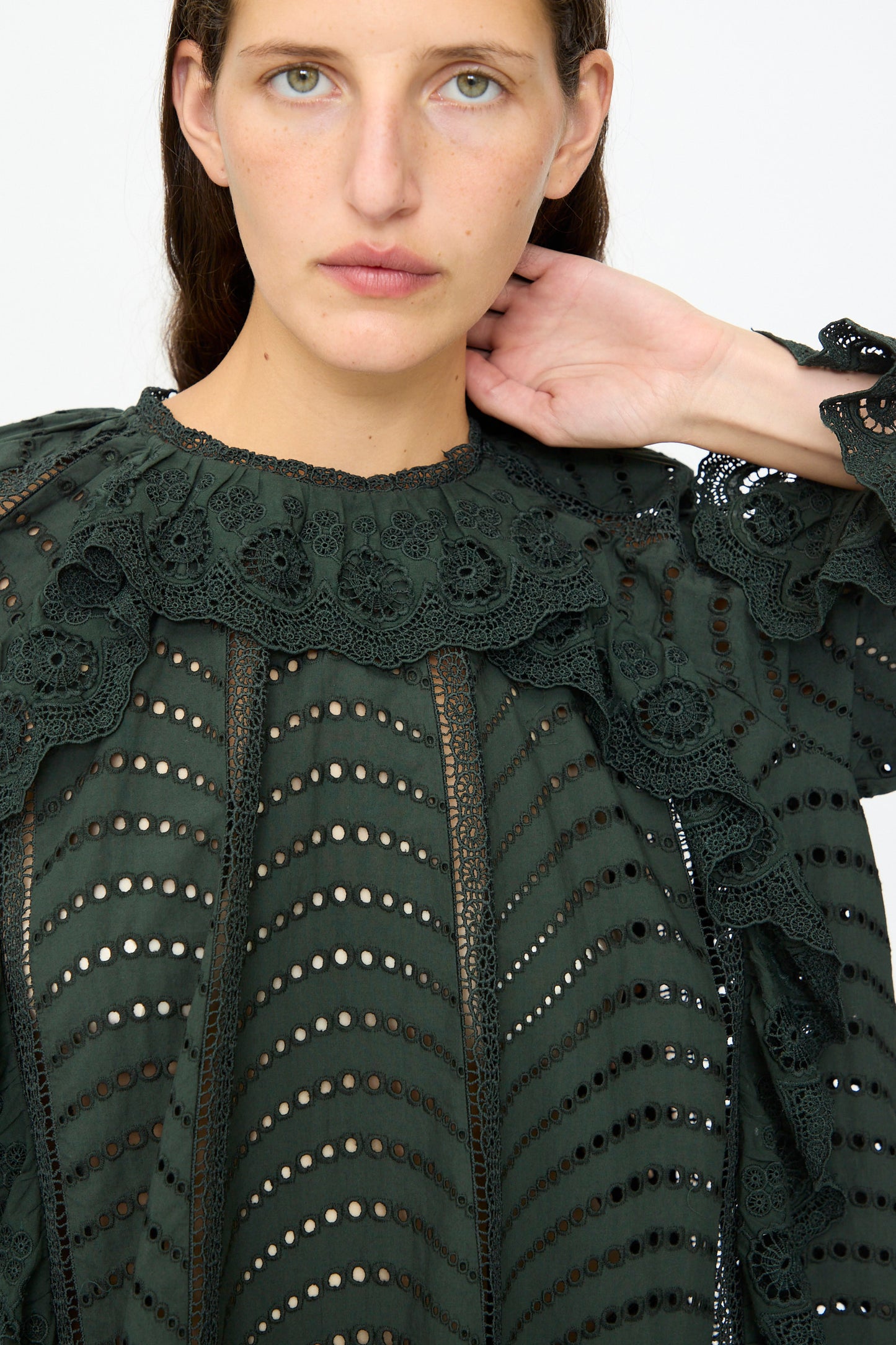 A person with light skin and dark hair wears the Ulla Johnson Marcella Blouse in Cypress, a dark green, long sleeve cotton top with eyelet lace and scalloped ruffles, facing the camera with a neutral expression.