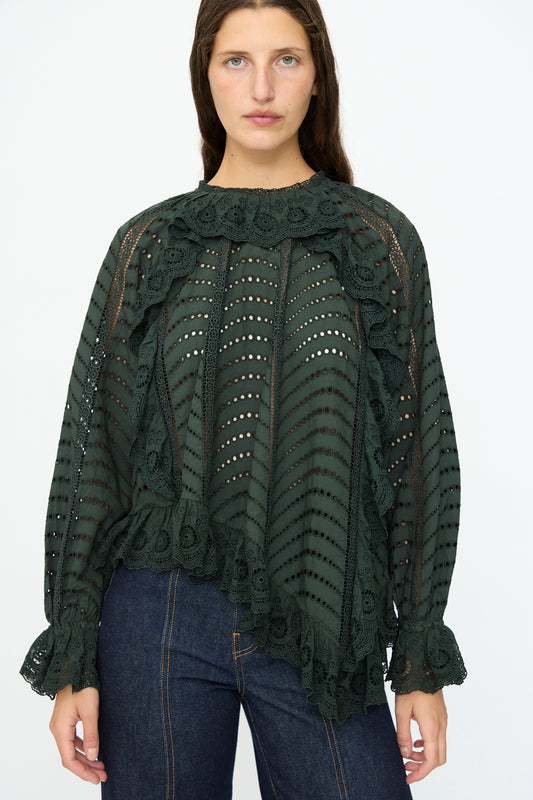 A woman wearing the Ulla Johnson Marcella Blouse in Cypress, featuring long sleeves, lace, and scalloped ruffles, paired with dark blue jeans against a plain white background.