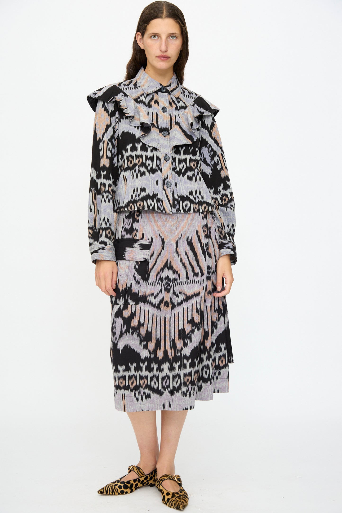A woman stands against a white background wearing the Ulla Johnson Mathilde Blouse in Tigereye with long sleeves, ikat motif, and ruffled shoulders, paired with leopard-print flat shoes.