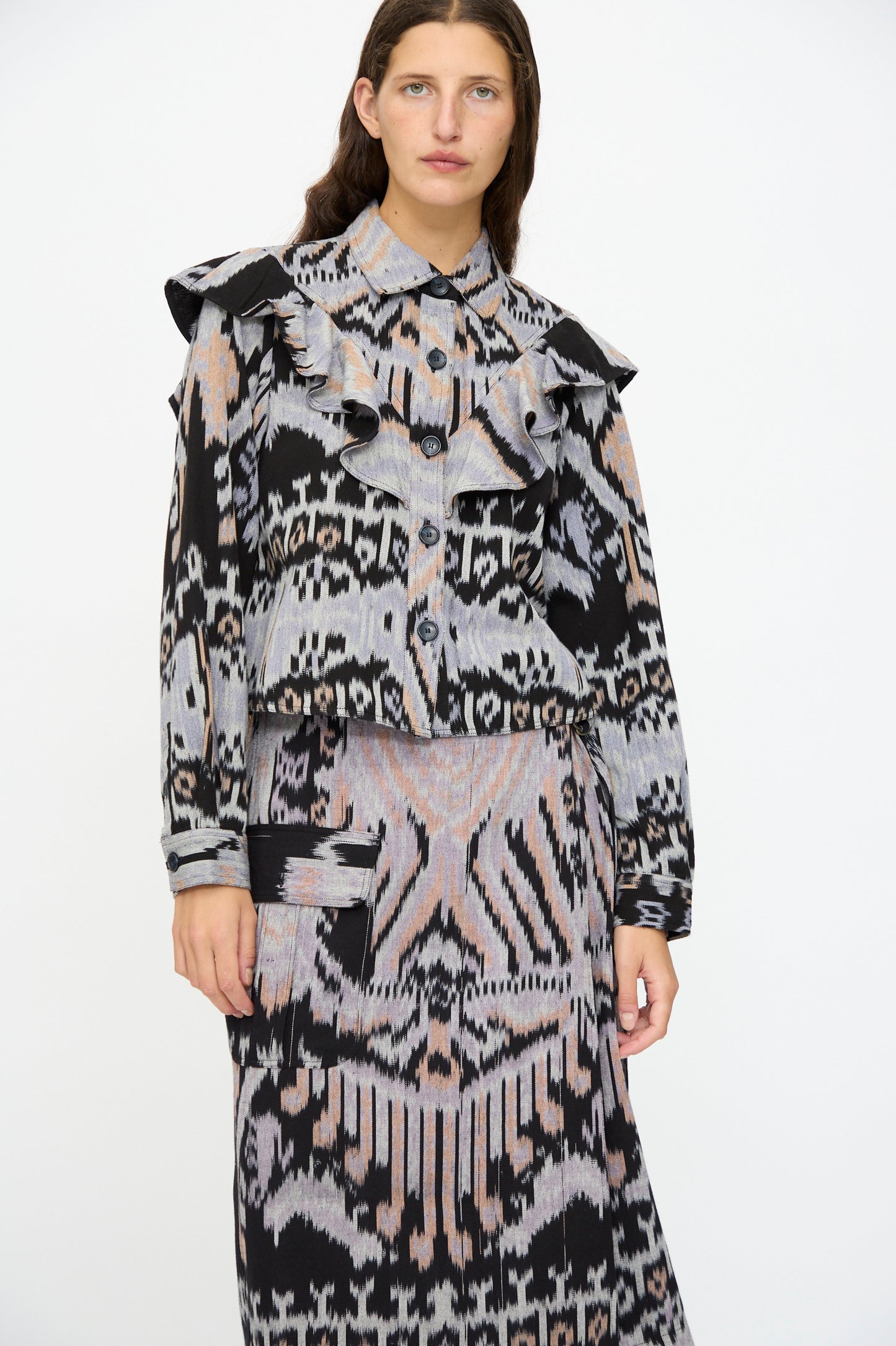 A woman wears the Ulla Johnson Mathilde Blouse in Tigereye, featuring long sleeves and ruffles, paired with a geometric skirt, standing against a plain white background.
