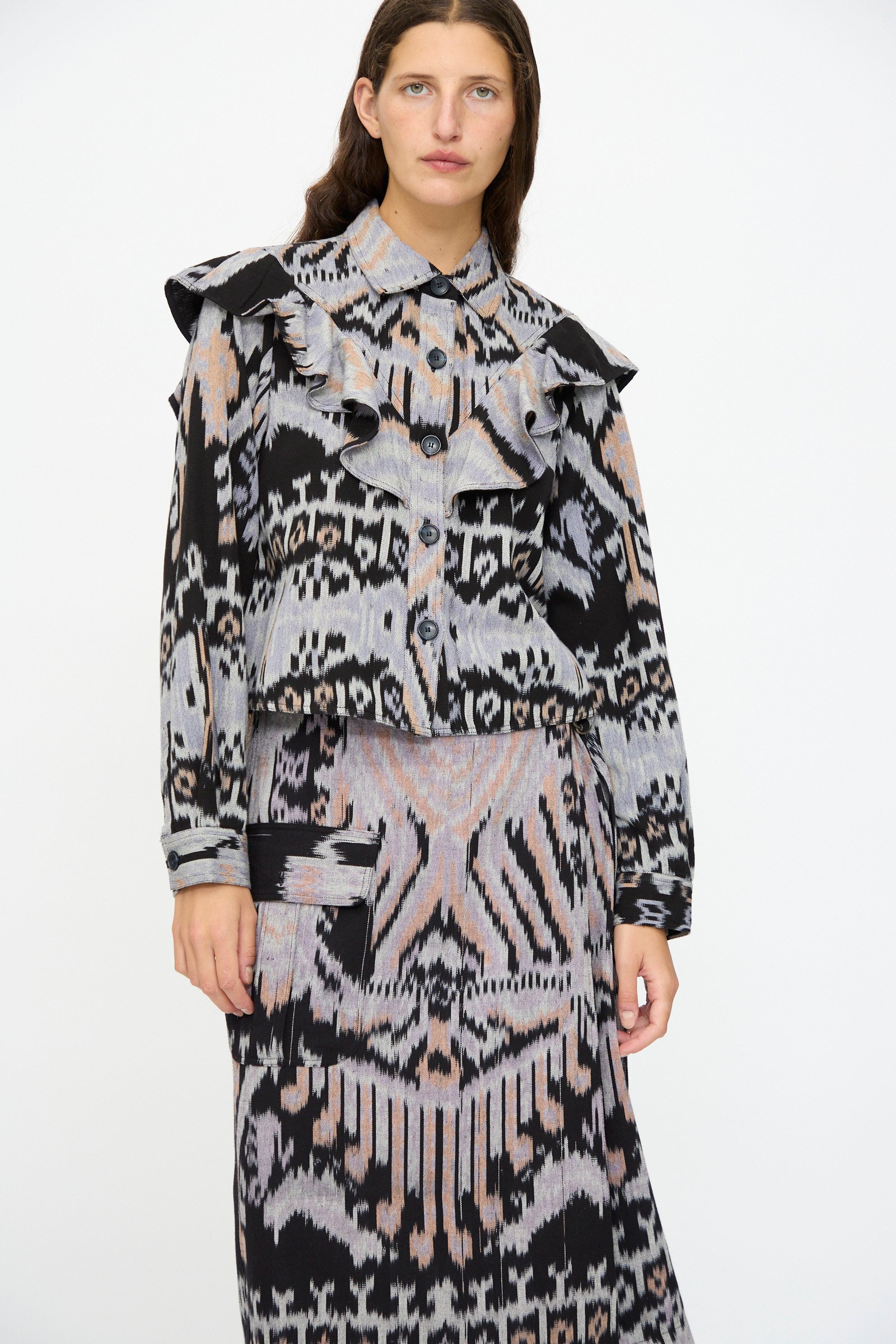 A woman wears the Ulla Johnson Mathilde Blouse in Tigereye, featuring long sleeves and ruffles, paired with a geometric skirt, standing against a plain white background.