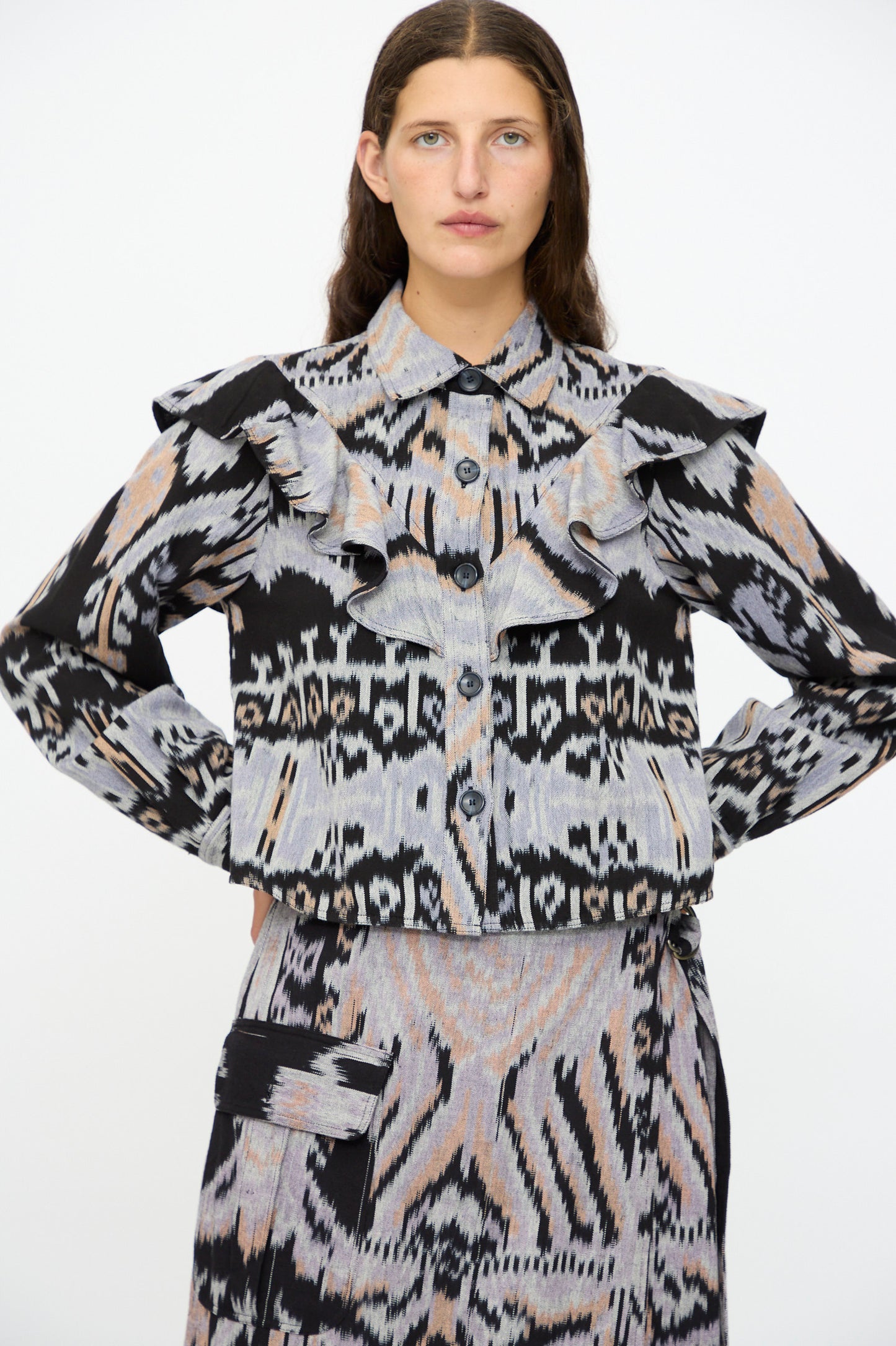 A woman poses with hands on hips, wearing the Ulla Johnson Mathilde Blouse in Tigereye—a patterned long sleeve cotton top with ruffles—paired with a matching skirt featuring pocket detail, set against a plain white background.