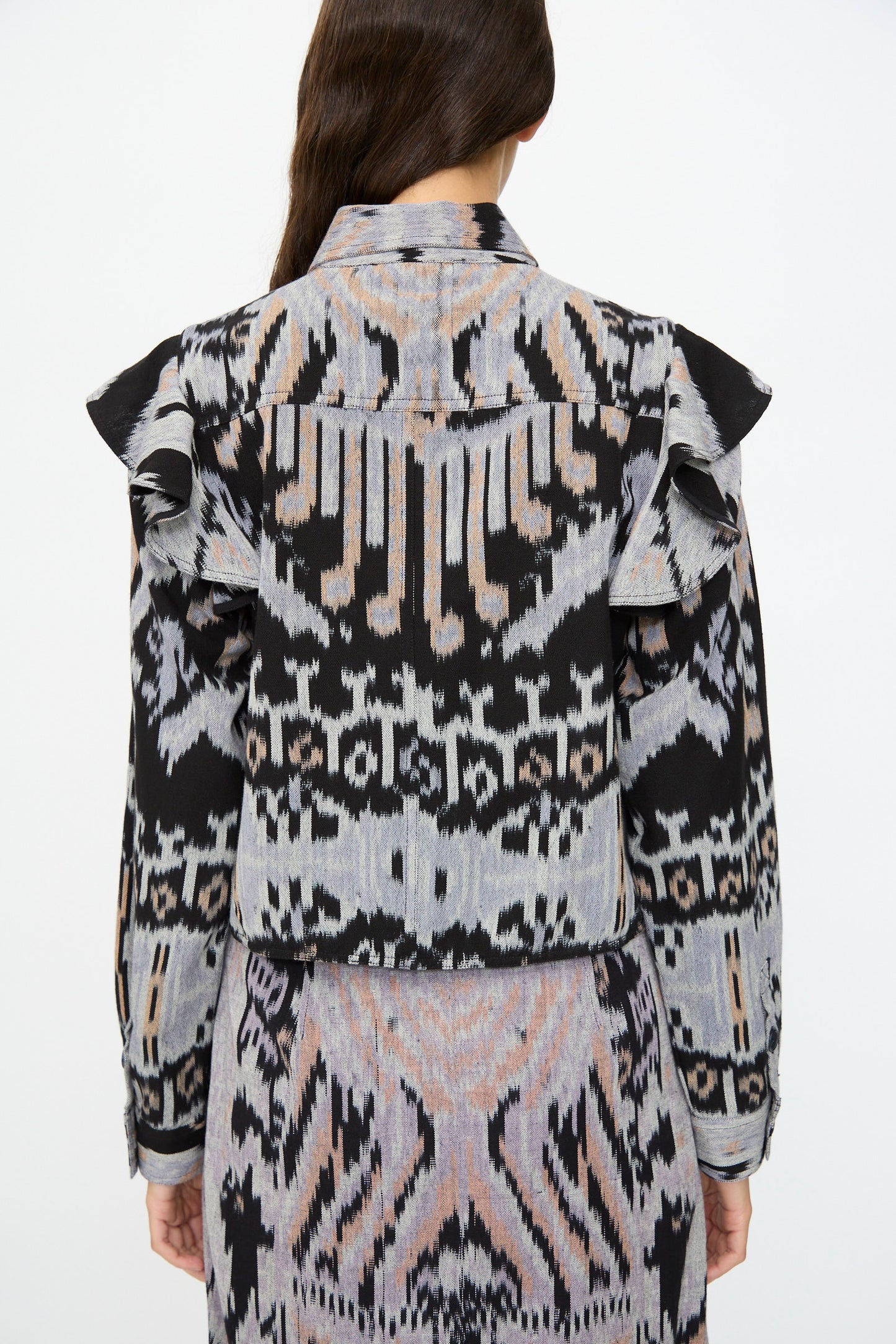 A person stands facing away, wearing the Ulla Johnson Mathilde Blouse in Tigereye with long sleeves, ruffled shoulders, and a matching patterned skirt.