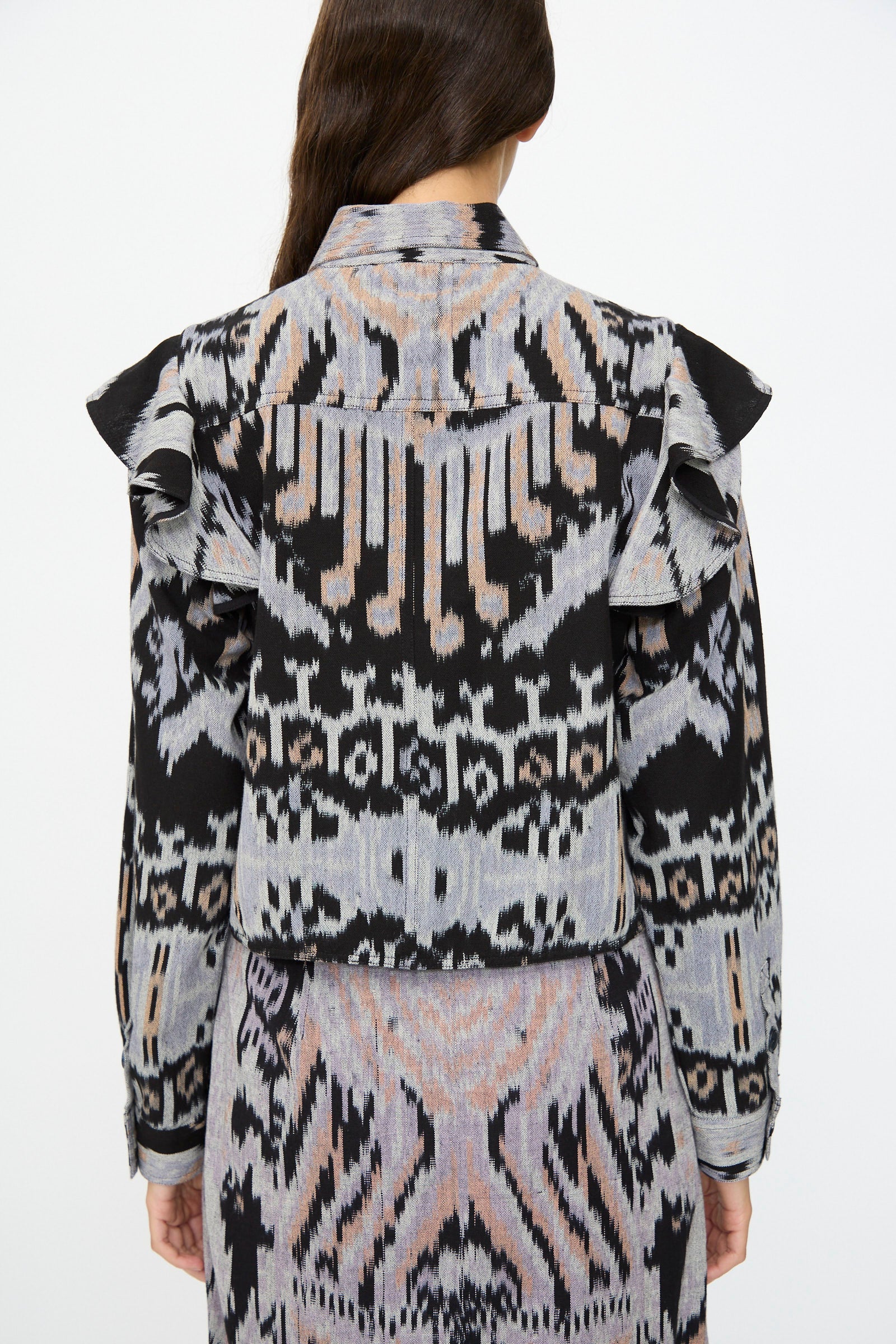 A person stands facing away, wearing the Ulla Johnson Mathilde Blouse in Tigereye with long sleeves, ruffled shoulders, and a matching patterned skirt.