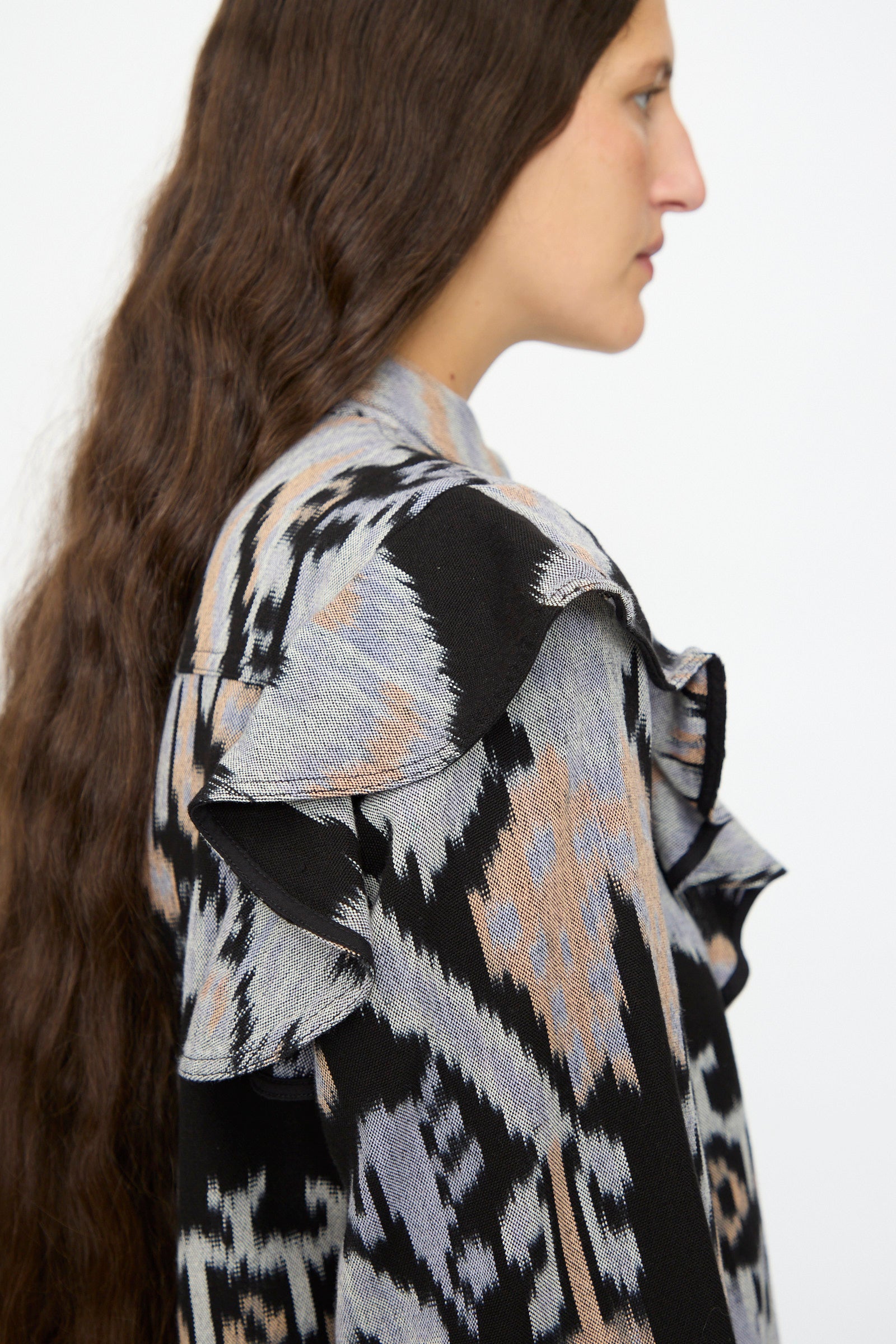 A woman with long, wavy brown hair wears the Ulla Johnson Mathilde Blouse in Tigereye, featuring hand-dyed cotton and ruffled shoulders, shown in profile against a plain background.