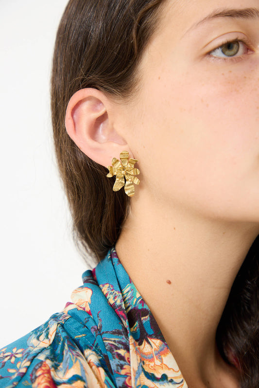 Close-up of a person's face wearing Ulla Johnson's Mini Floret Stud in Brass and a multicolored patterned shirt. The person has long brown hair and light skin.