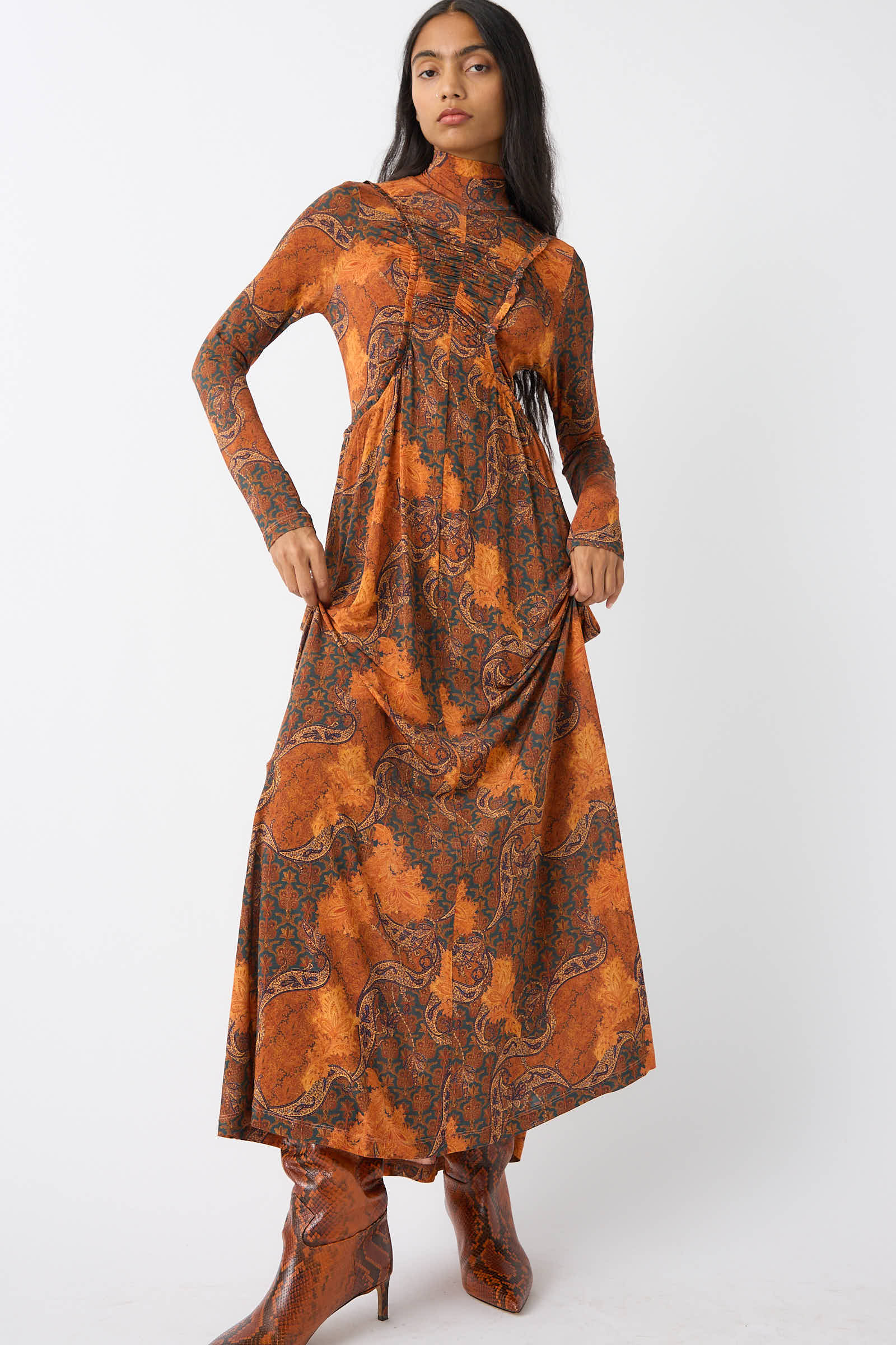 A person wearing the Ulla Johnson Oriana Dress in Amber, which features a vibrant paisley print, and matching orange boots stands against a plain white background.