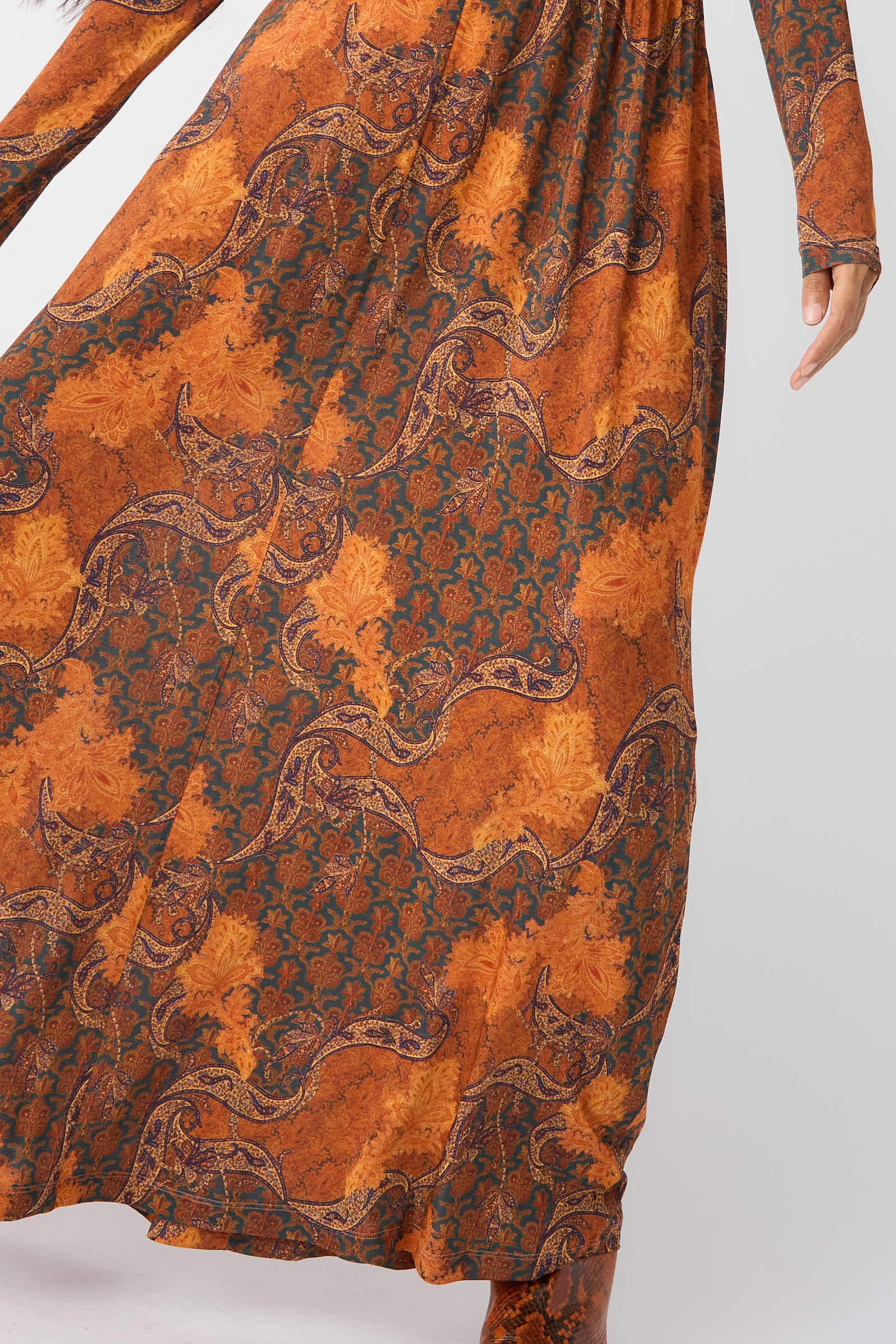 A person wearing the Oriana Dress in Amber by Ulla Johnson, extending their arm to the side. The silken tricot jersey dress features intricate floral and ornate patterns in vibrant orange and green paisley print.