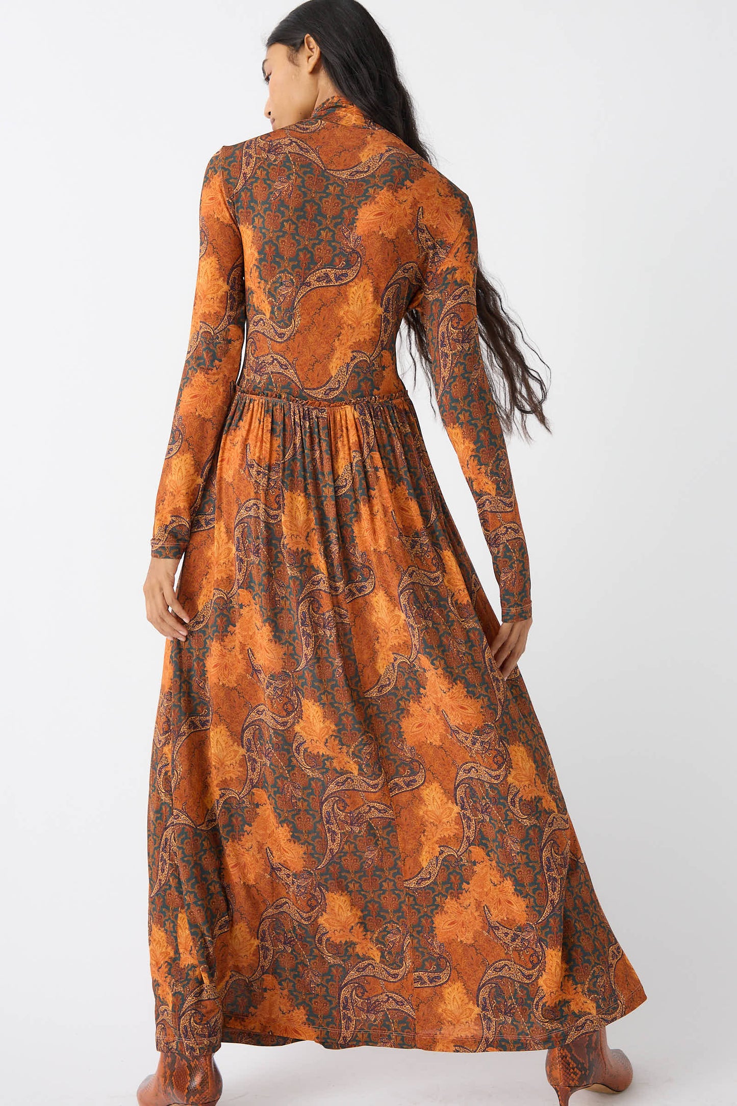 A person wearing the Oriana Dress in Amber by Ulla Johnson, a long-sleeve, floor-length garment with an orange and green patterned design, stands facing away to showcase its vibrant paisley print against a plain white background.