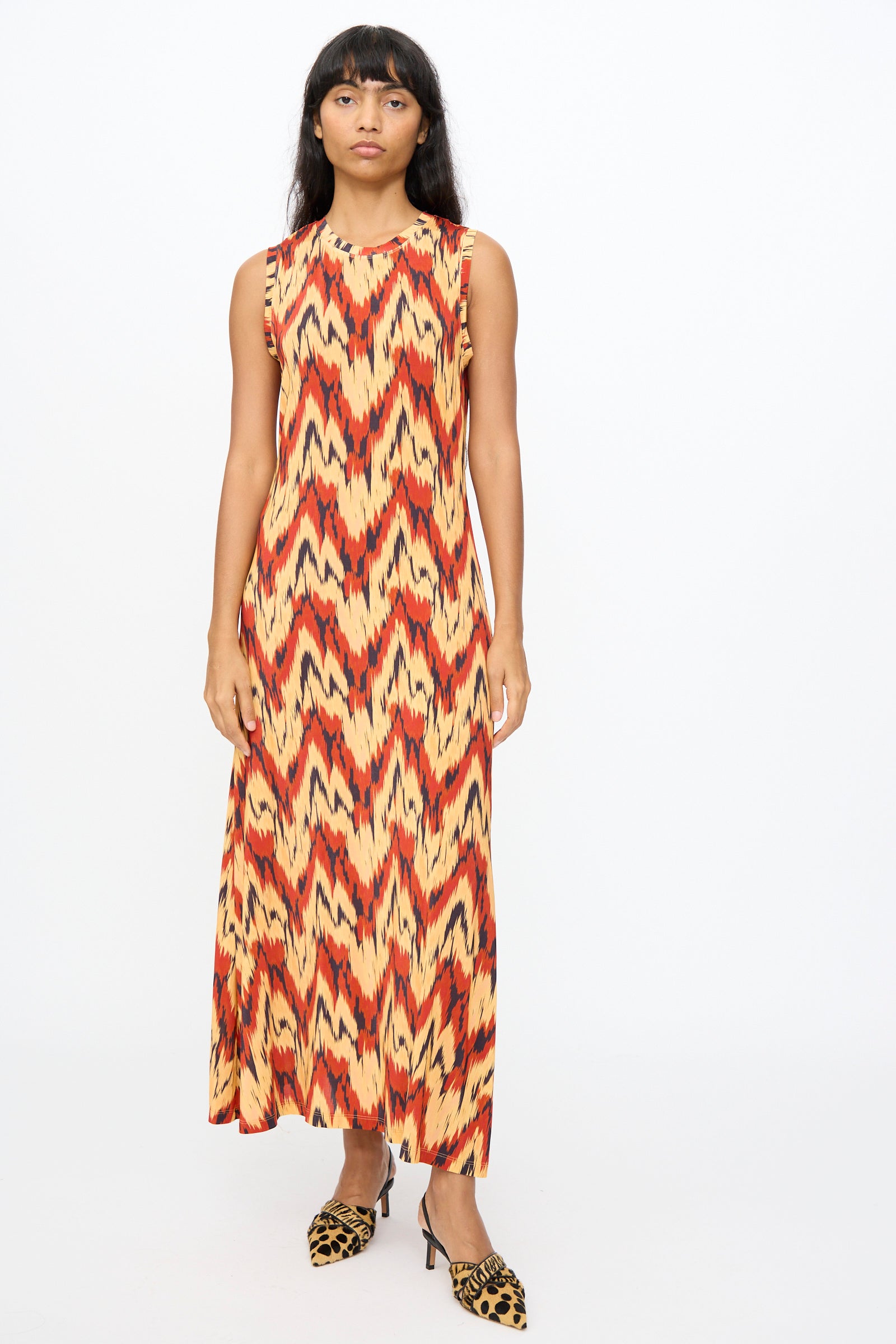 A woman faces forward wearing the Ulla Johnson Orla Dress in Carnelian, a sleeveless tricot jersey dress with bold red, orange, and yellow zigzag patterns, paired with animal print shoes.