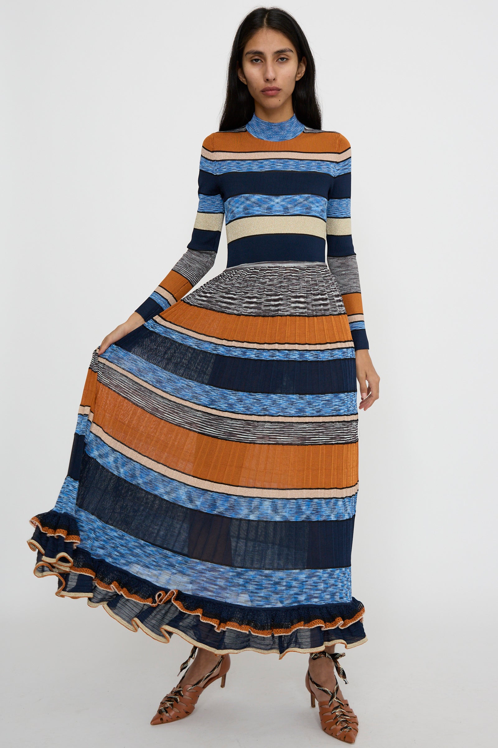 A person stands forward wearing the Pleated Striped Knit Evangeline Dress in Adriatic by Ulla Johnson, featuring a long, multicolored maxi design with blue, orange, and gray patterns.