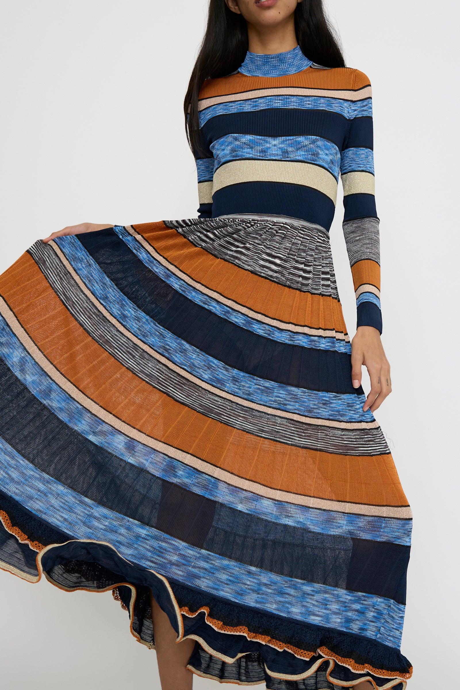 A person wearing the Pleated Striped Knit Evangeline Dress in Adriatic by Ulla Johnson showcases the space-dyed stripes with blue, orange, and black horizontal patterns as they lift the skirt to the side. 