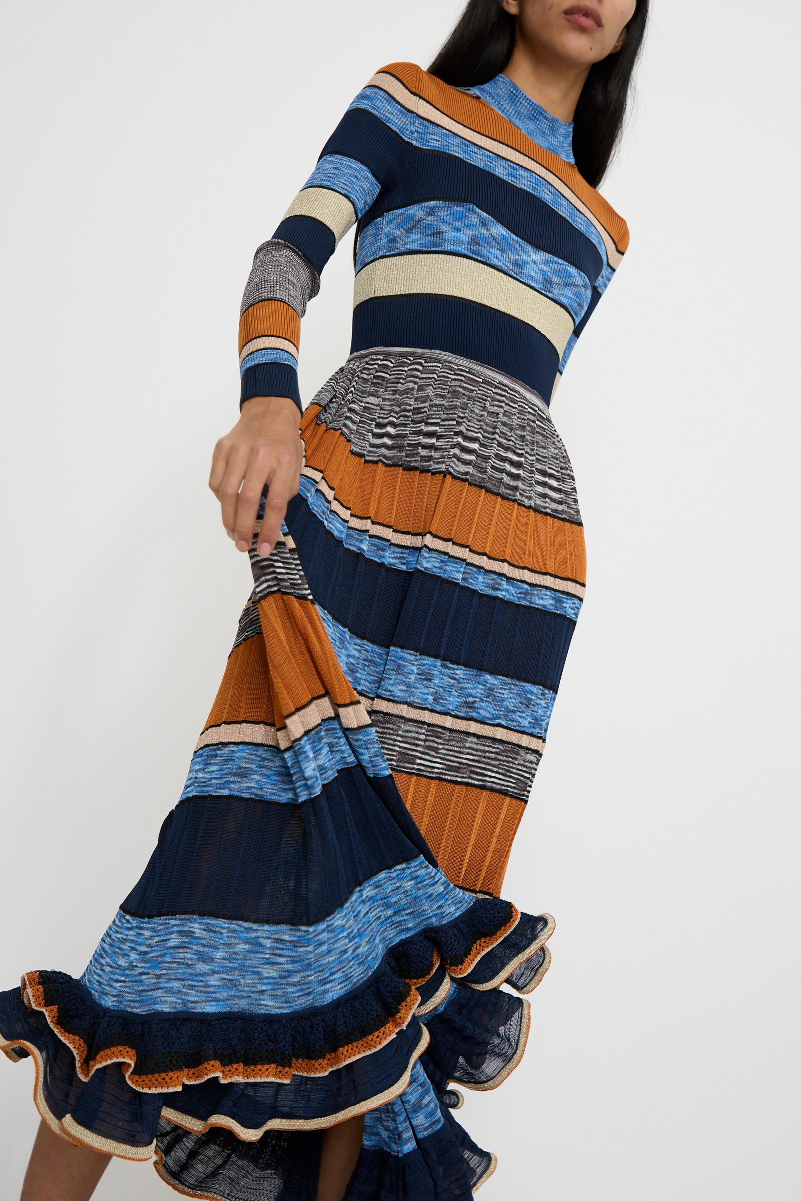 A person stands against a white background, wearing the Pleated Striped Knit Evangeline Dress in Adriatic by Ulla Johnson. The dress features long sleeves and showcases a space-dyed design with a mix of blue, orange, and gray patterns. 