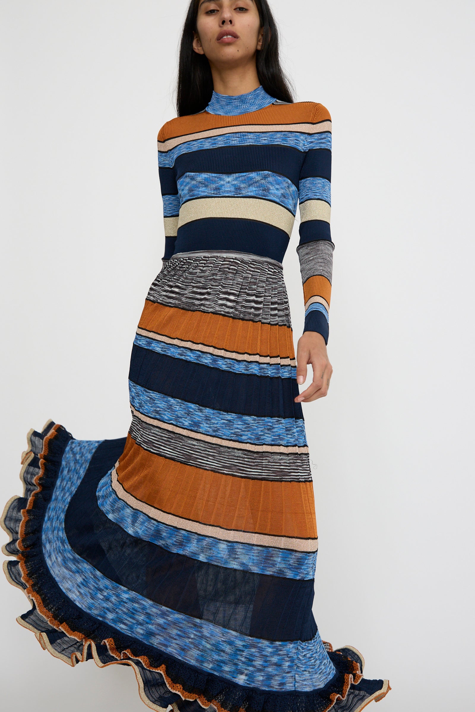 A person is wearing the Pleated Striped Knit Evangeline Dress in Adriatic by Ulla Johnson, a long-sleeve maxi dress with a flowing skirt, showcasing shades of blue, orange, and beige, against a white background. 