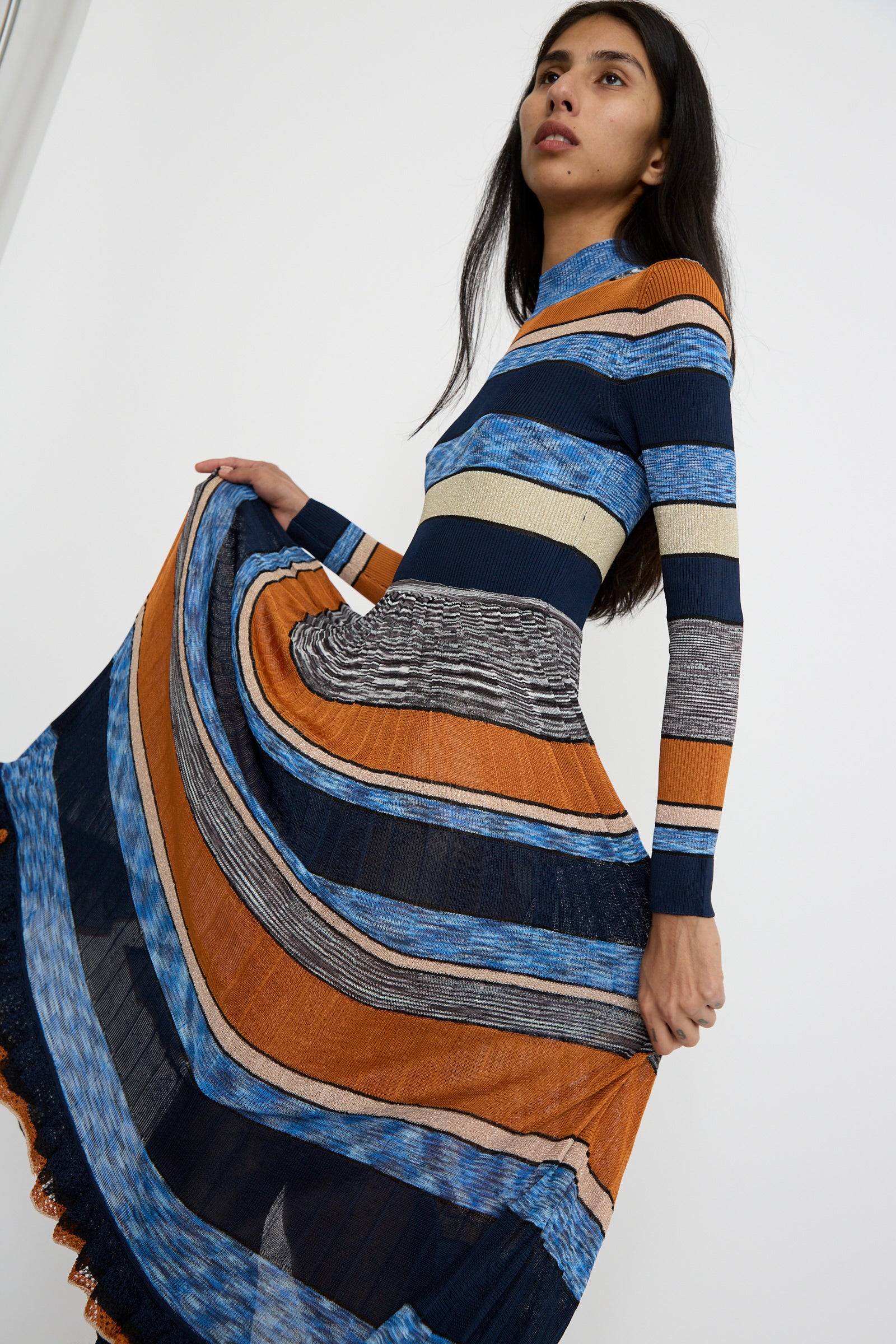 A person elegantly holds out the skirt of the Pleated Striped Knit Evangeline Dress in Adriatic by Ulla Johnson, showcasing its vibrant orange, blue, and black stripes against a white background. 