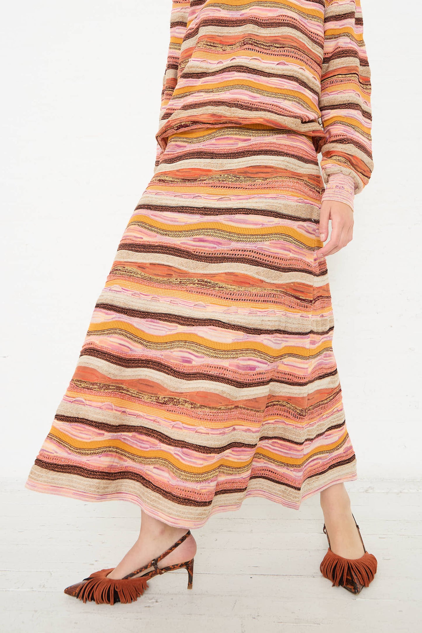 A person wearing a patterned, long-sleeved top and the Rosen Skirt in Sunset by Ulla Johnson, a jacquard knit maxi skirt with an elasticated waistband in various shades of pink, orange, and brown. The ensemble features wavy stripes and is complemented by brown fringed footwear.