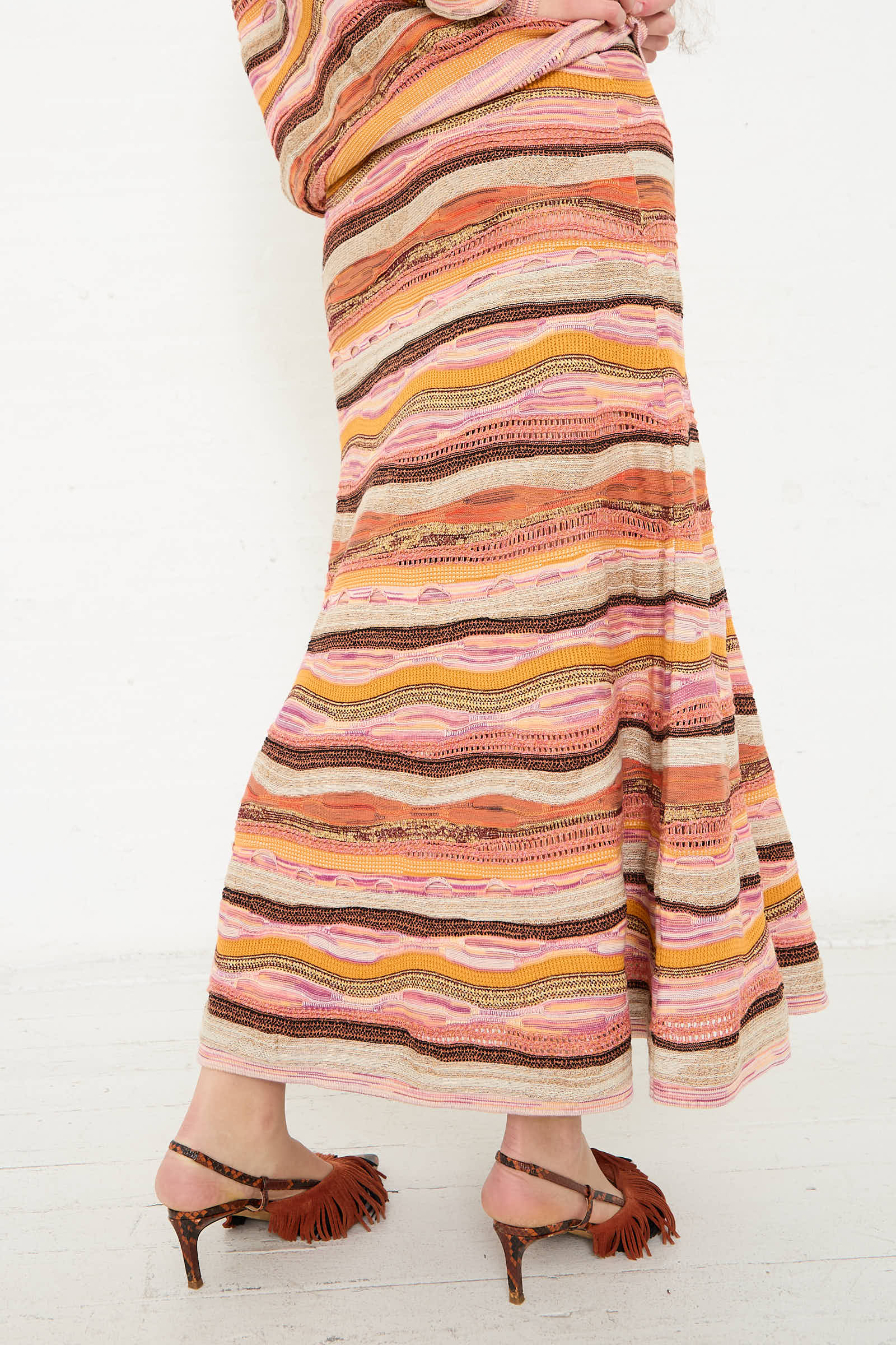 Close-up of a person wearing the Ulla Johnson Rosen Skirt in Sunset, a colorful jacquard knit maxi skirt with wavy stripes, and brown high-heeled sandals with fringe details, standing against a white background.