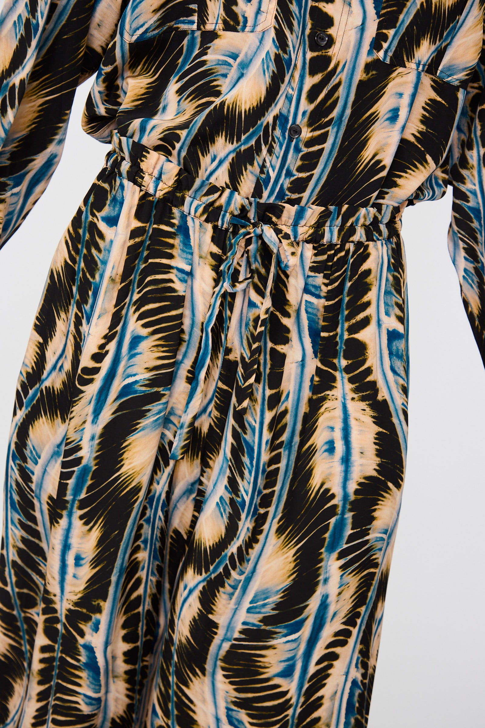 Close-up of a person wearing Ulla Johnson's Shibori Wave Printed Sawyer Pant in River, featuring black, tan, and blue abstract silk with a gathered waist and matching long-sleeve shirt.