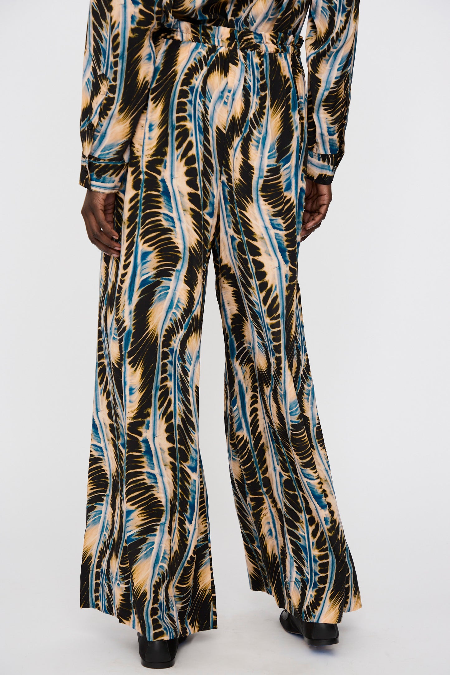 A person wears the Ulla Johnson Shibori Wave Printed Sawyer Pant in River—oversized, wide-leg silk pants with an abstract feather-like pattern—paired with a matching top and black shoes.