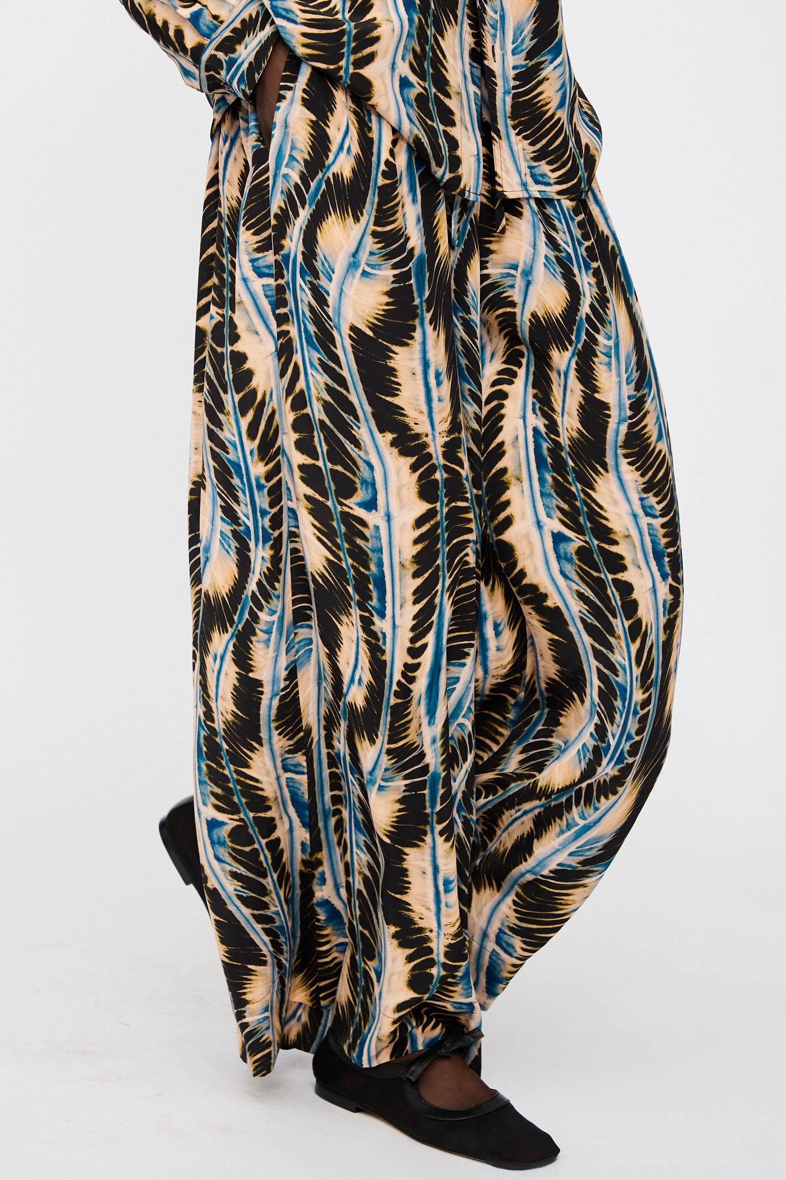 Person wearing the Ulla Johnson Shibori Wave Printed Sawyer Pant in River, featuring a black, beige, blue, and white abstract pattern, paired with black slip-on shoes against a plain background.