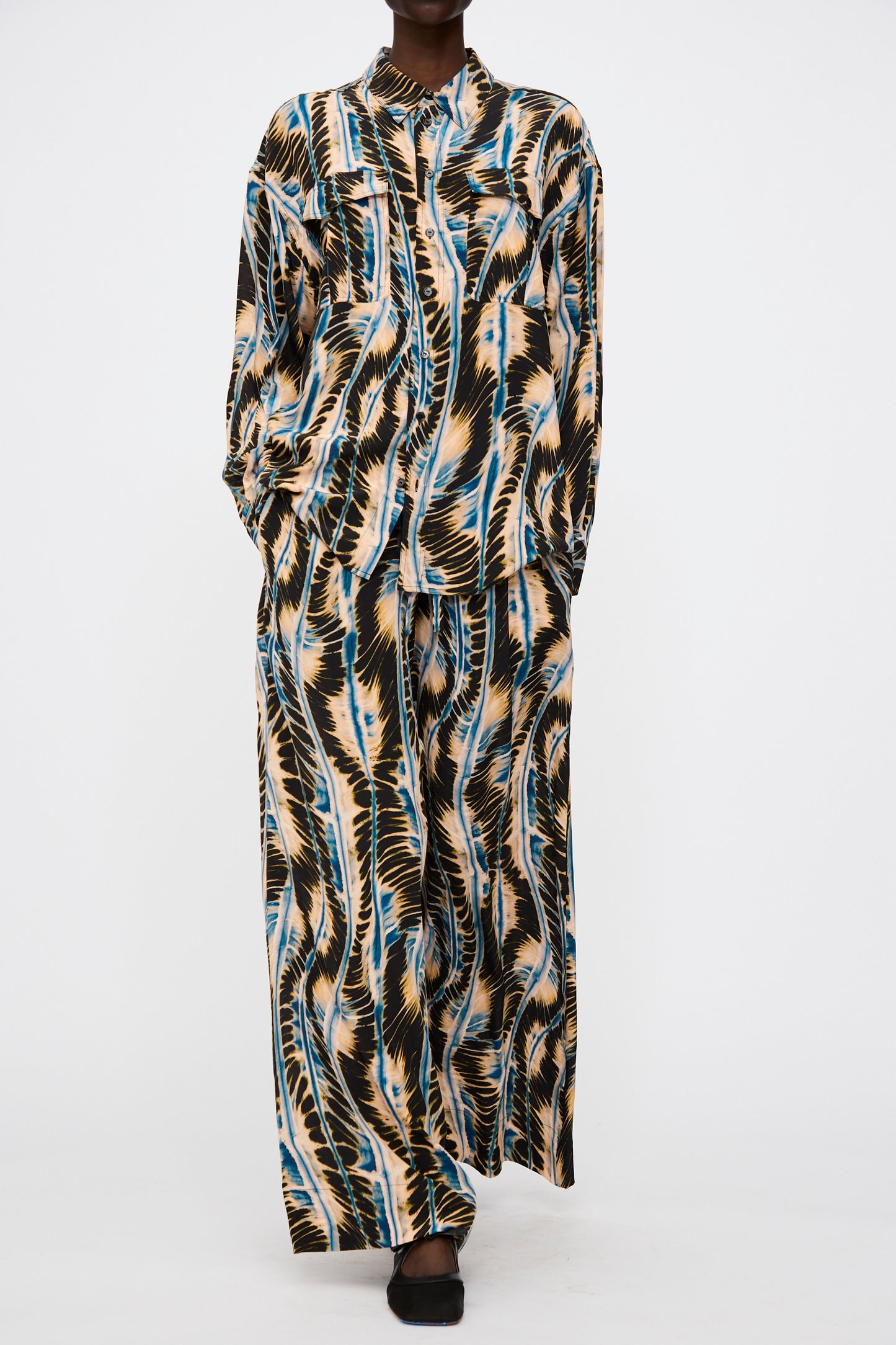 Model wears the Ulla Johnson Shibori Wave Printed Sawyer Pant in River—a silk, abstract-patterned oversized pant set—paired with a matching long-sleeve button-up shirt, shown against a plain white background.