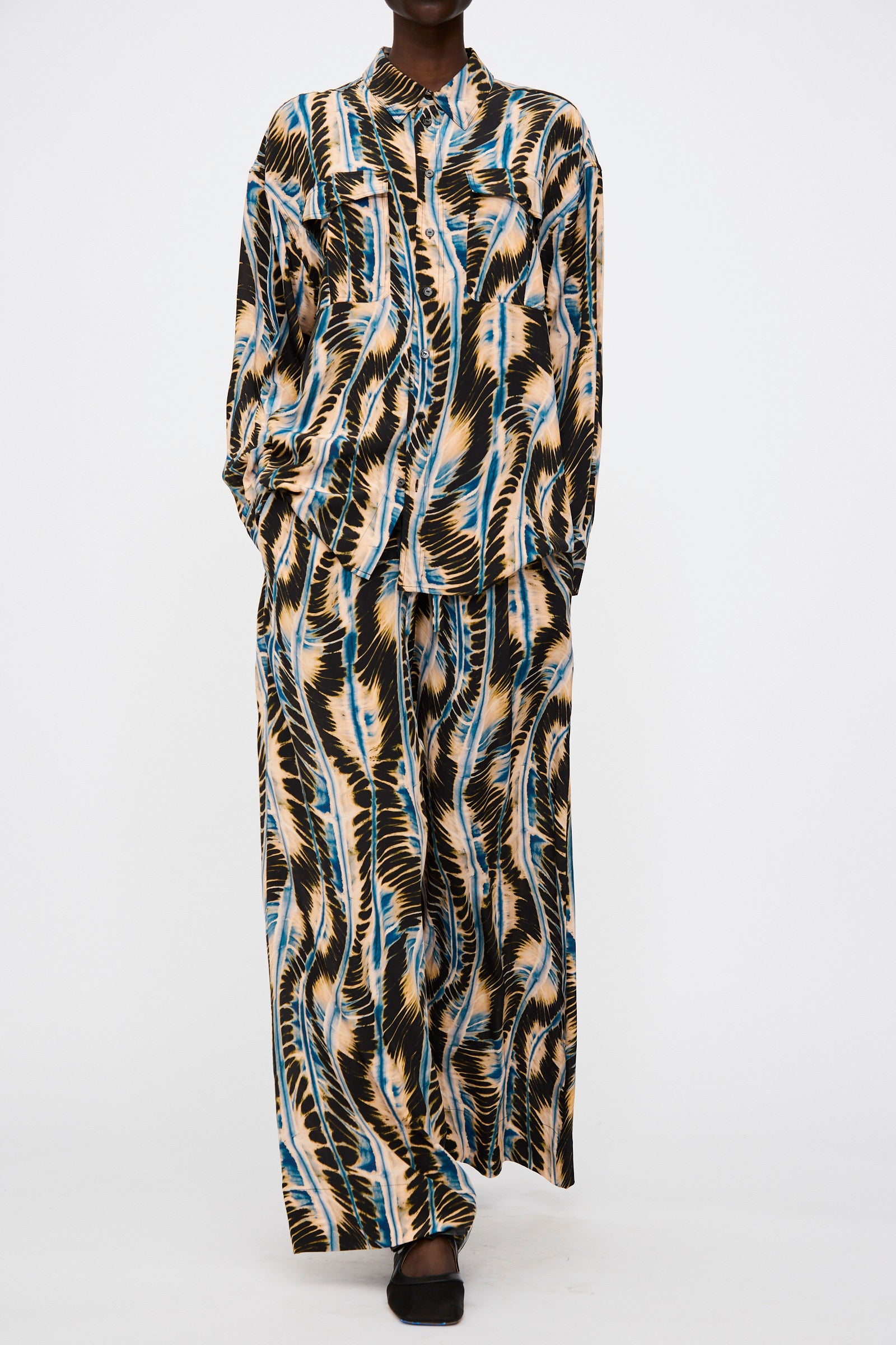 Model wears the Ulla Johnson Shibori Wave Printed Sawyer Pant in River—a silk, abstract-patterned oversized pant set—paired with a matching long-sleeve button-up shirt, shown against a plain white background.