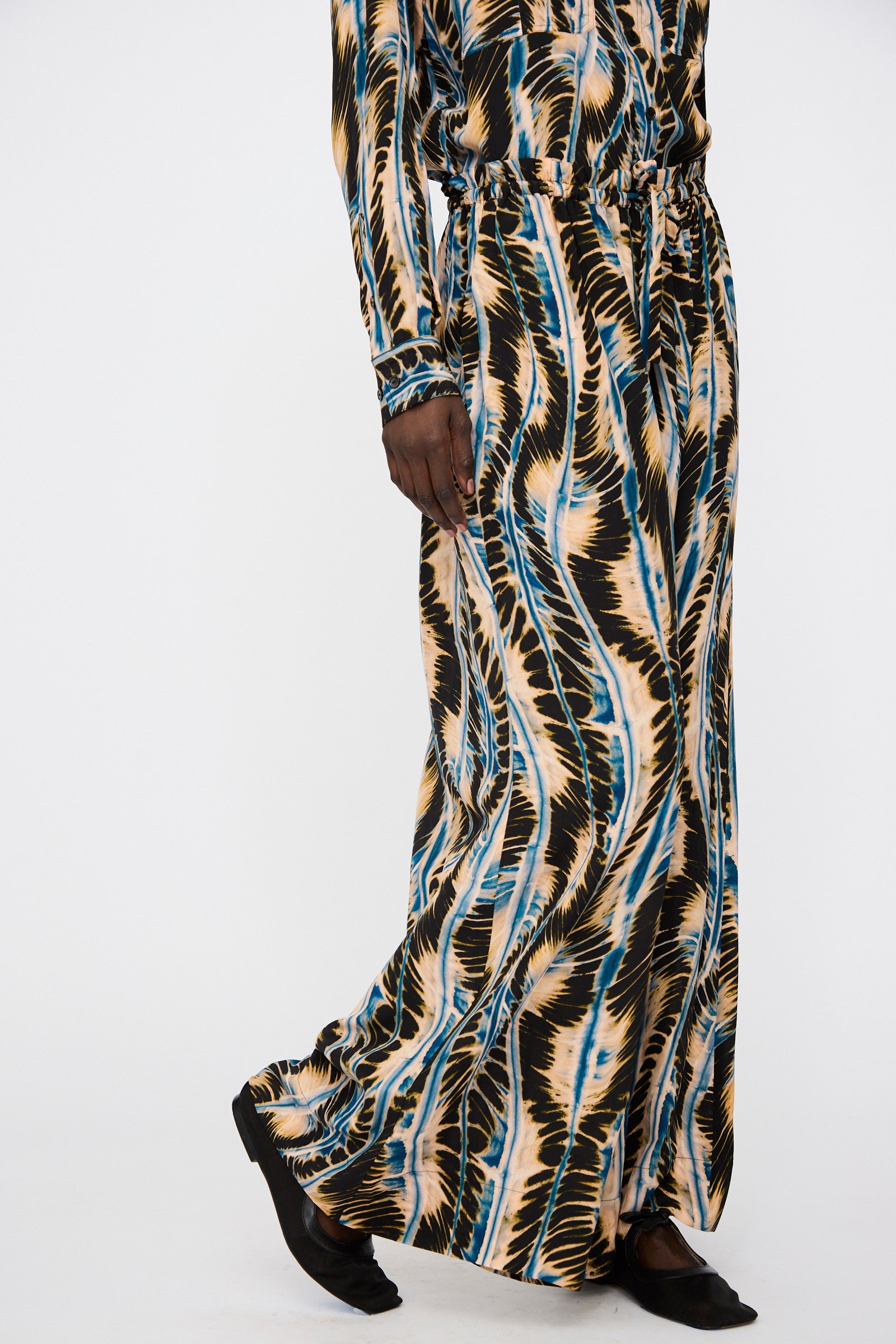 Model wears the Ulla Johnson Shibori Wave Printed Sawyer Pant in River, featuring black, beige, and blue zigzag stripes, paired with black shoes against a plain white background.