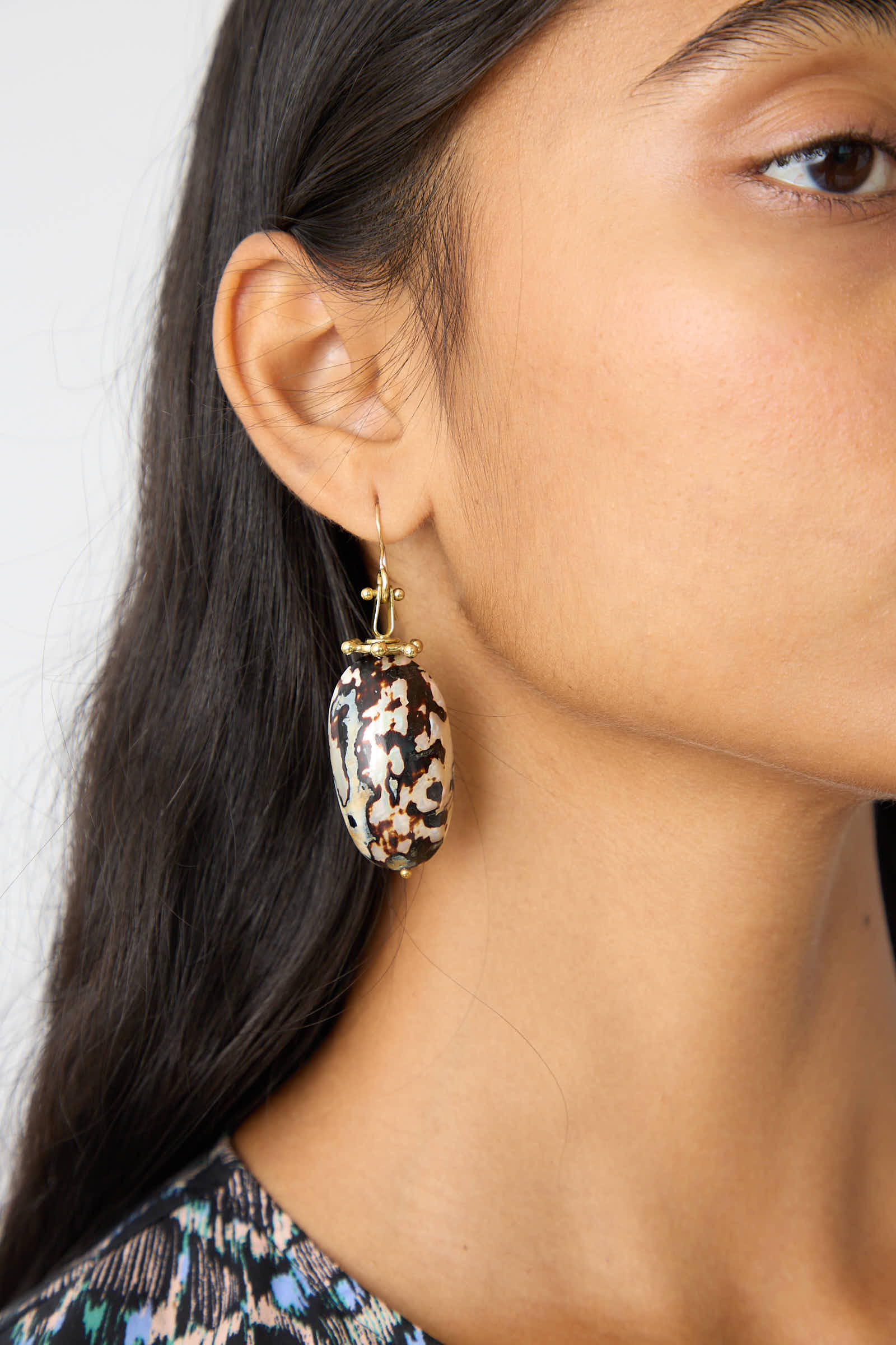 Close-up of a person wearing the Silayah Earring in Shell by Ulla Johnson, a large, oval-shaped handcrafted accessory showcasing a black, brown, and white pattern attached to a gold hook. The person's long dark hair is worn down. This piece draws inspiration from Byzantine jewelry, adding a touch of ancient elegance.