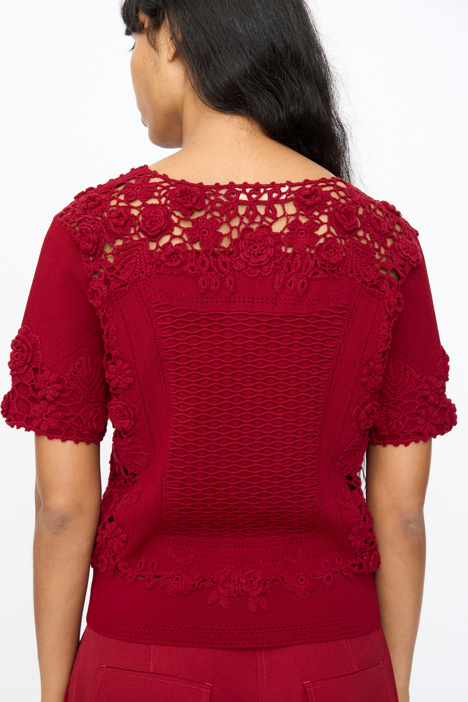 A woman is seen from the back wearing the Ulla Johnson Stella Knit Top in Begonia, a short sleeve style with intricate crochet and lace details on the shoulders, sleeves, and upper back, finished with a scalloped hem.