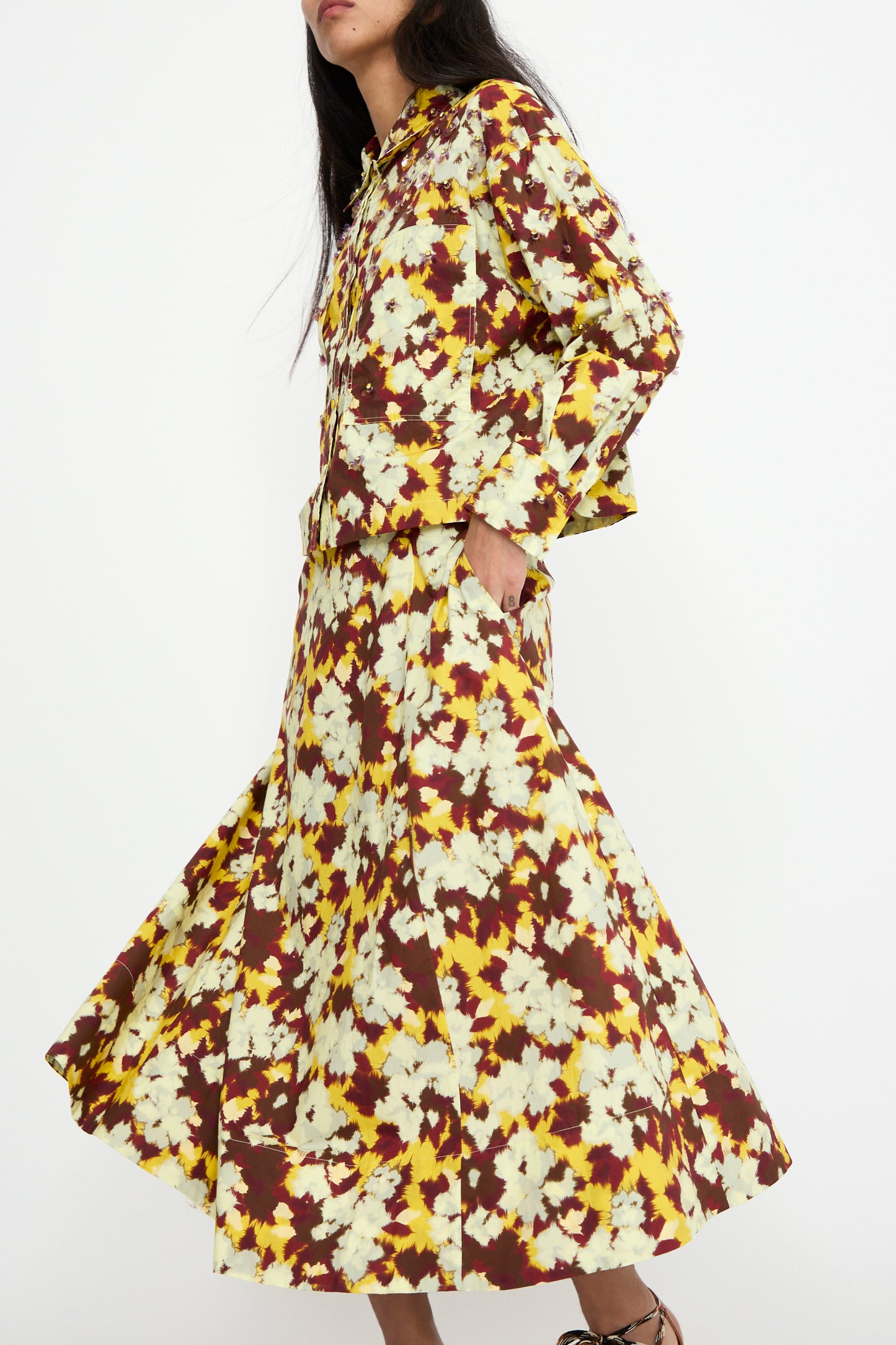 A person wearing a floral-patterned blouse in shades of brown, yellow, and white weara it with the Vianne Skirt in Acacia by Ulla Johnson, placing their hands in the skirt's pockets against a plain background.