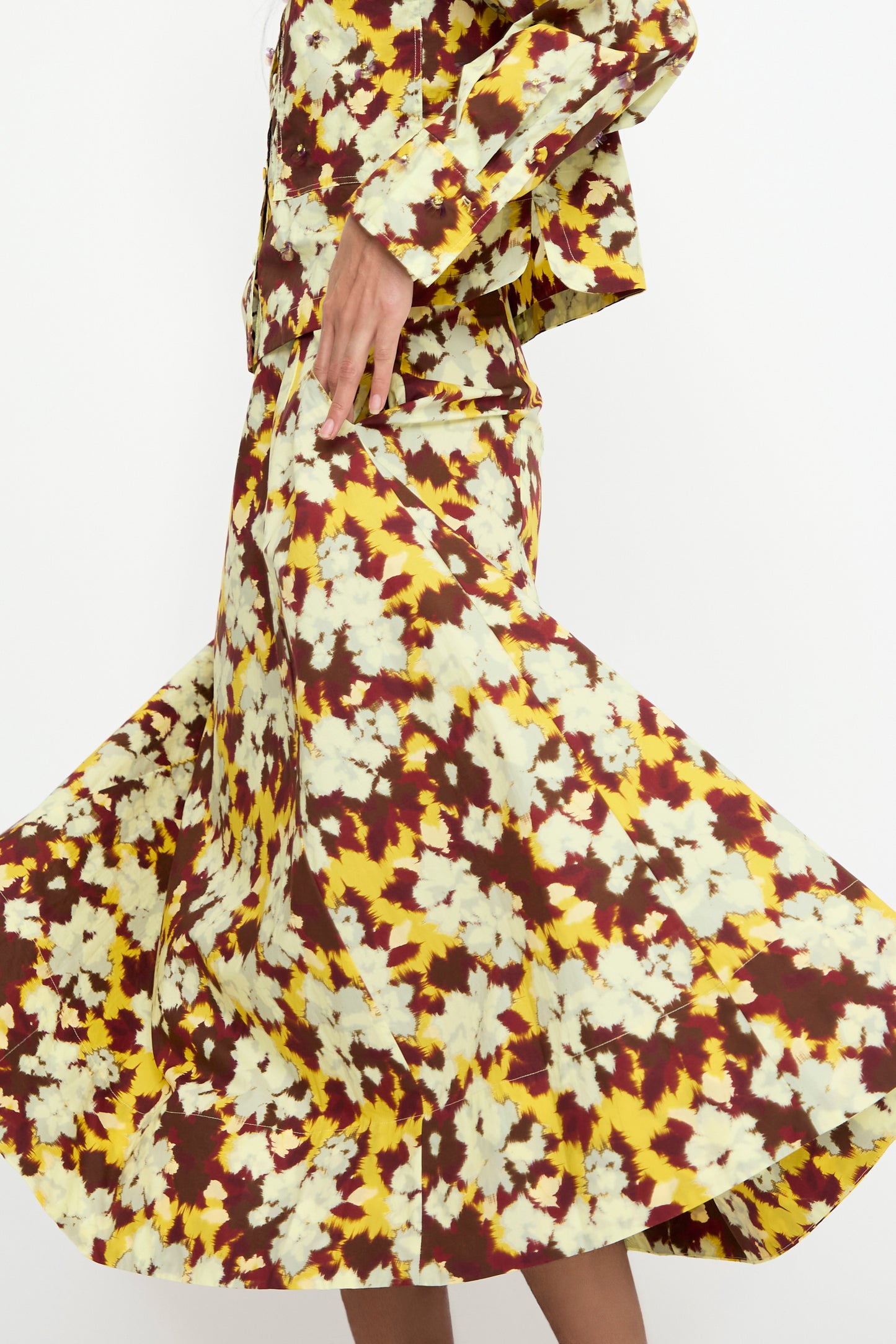 Wearing the Vianne Skirt in Acacia by Ulla Johnson, a vibrant yellow, white, and maroon floral-patterned ensemble, the person is captured mid-movement, twirling their A-line midi skirt made from soft cotton poplin.