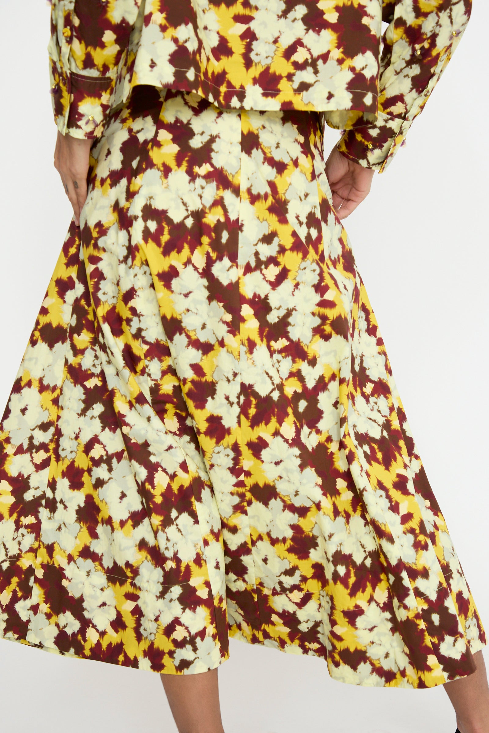 A person is wearing the Vianne Skirt in Acacia by Ulla Johnson, along with a matching top, both crafted from cotton poplin and showcasing a vibrant ikat floral pattern in yellow, brown, and white.